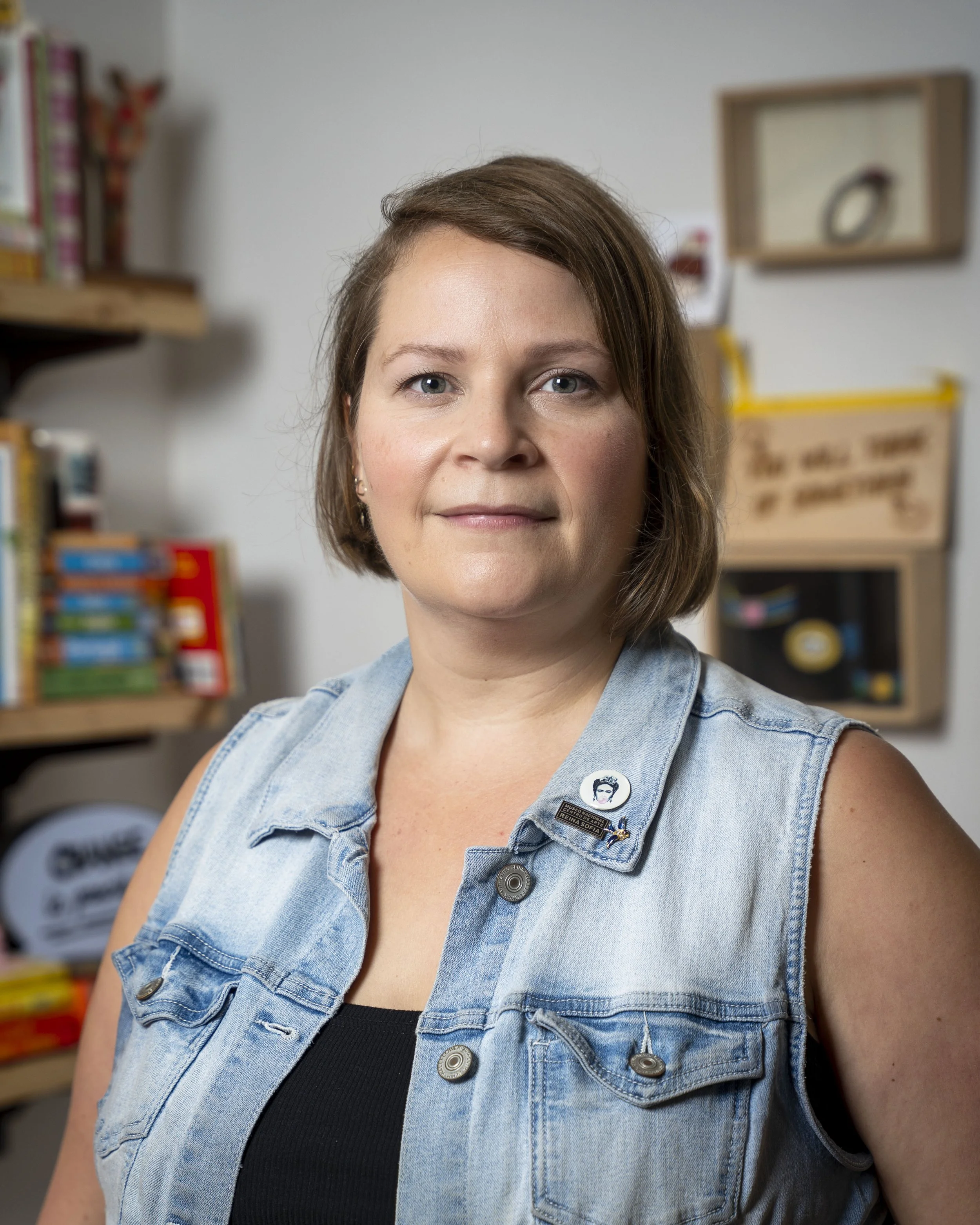 Close-up of a woman with shoulder-length brown hair and blue eyes, wearing a sleeveless denim vest with pins and a black top underneath, standing in a room with bookshelves and a sign in the background.