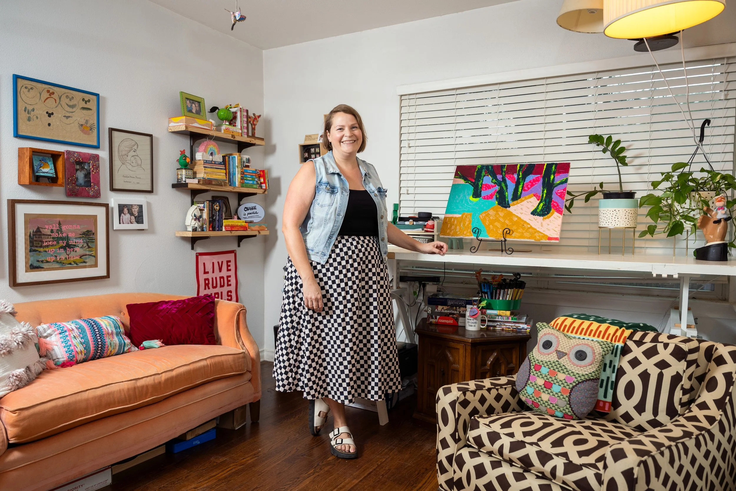 A woman smiling and standing in a cozy, decorated room with artwork, books, and colorful cushions on a sofa.