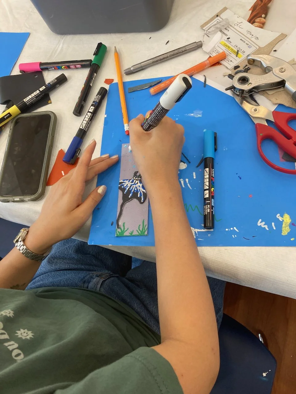 Person using paint pens to decorate a handmade leather bookmark at a Kiki Knows Art workshop.