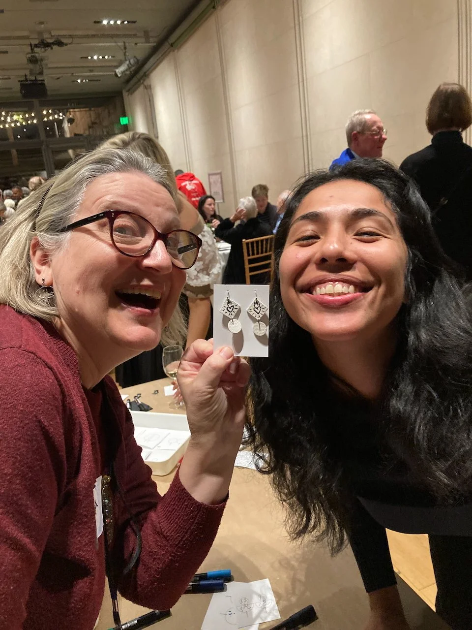 Two smiling women holding a pair of white earrings made at a Kiki Knows Art jewelry workshop.