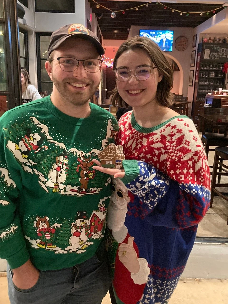 A man and woman smiling and holding their handmade gingerbread house ornament at a Kiki Knows Art workshop.