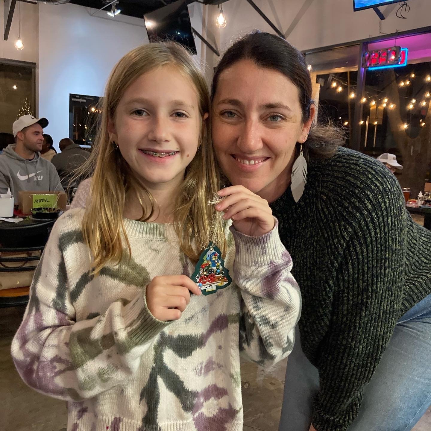 A young girl and woman smiling at a Kiki Knows Art workshop, holding a handmade Christmas ornament.