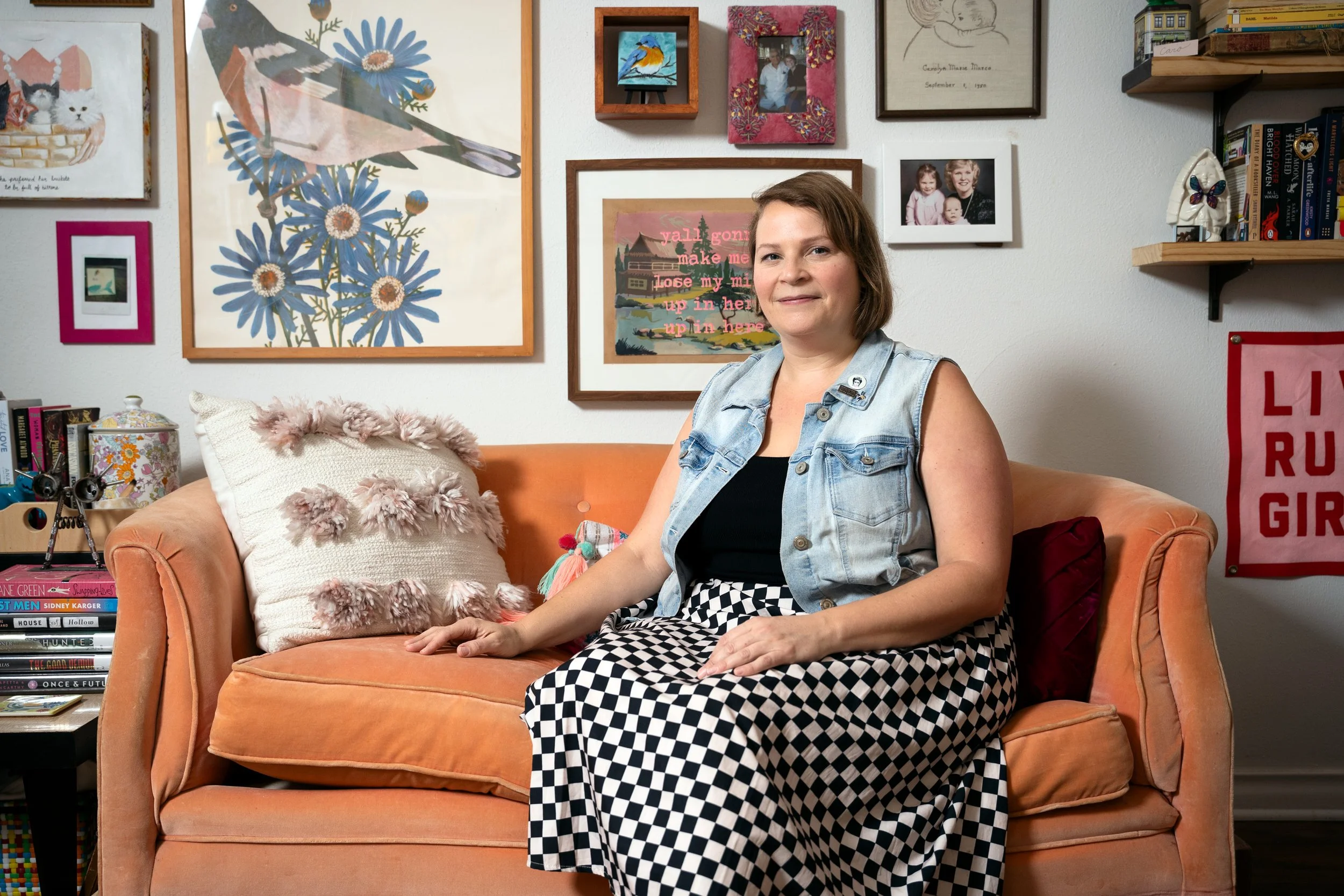 A woman sitting on an orange couch in a room decorated with various framed pictures, paintings, and shelves. She has short brown hair, wears a denim vest over a black top, and a black and white checkered skirt. Behind her are books, a pink pillow with pom-pom trim, and eclectic artwork on the wall.