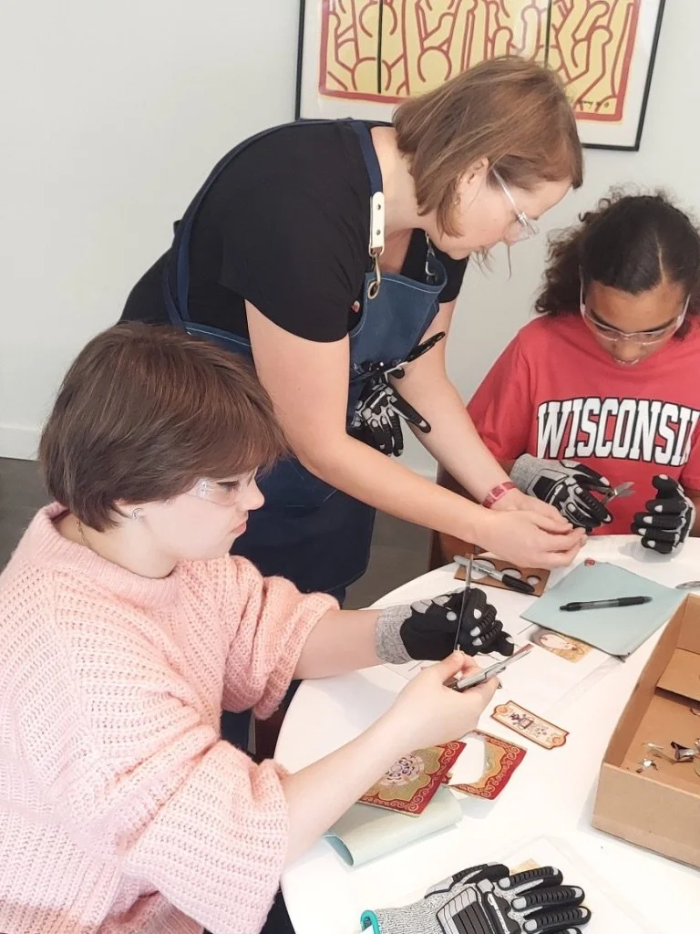 A woman named Carolyn Marco assisting two children with a jewelry-making activity at a table, with tools, jewelry pieces, and colorful envelopes, in a room with framed artwork on the wall.
