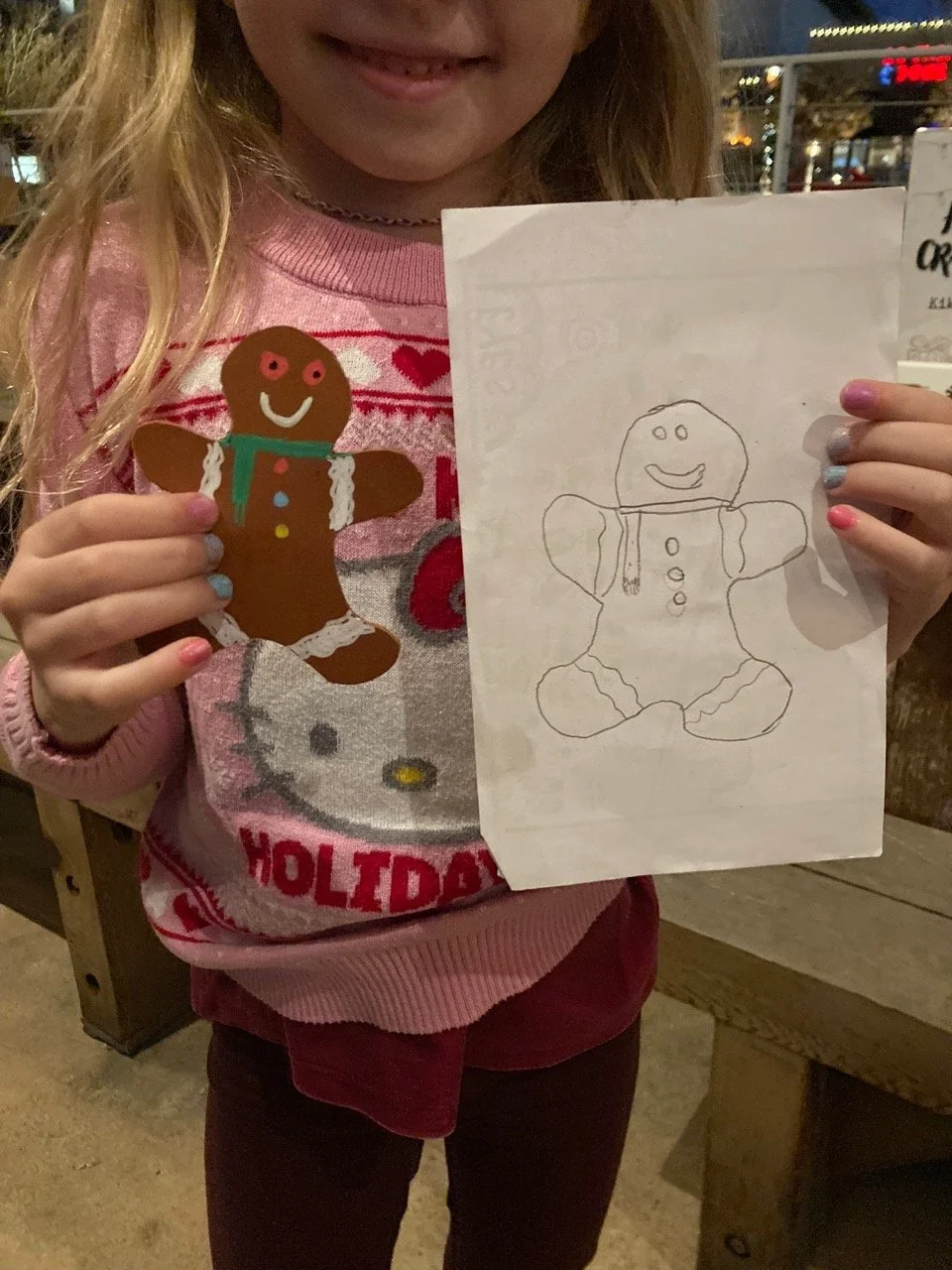 A young girl holding her handmade gingerbread man alongside her drawing of it at a Kiki Knows Art workshop.