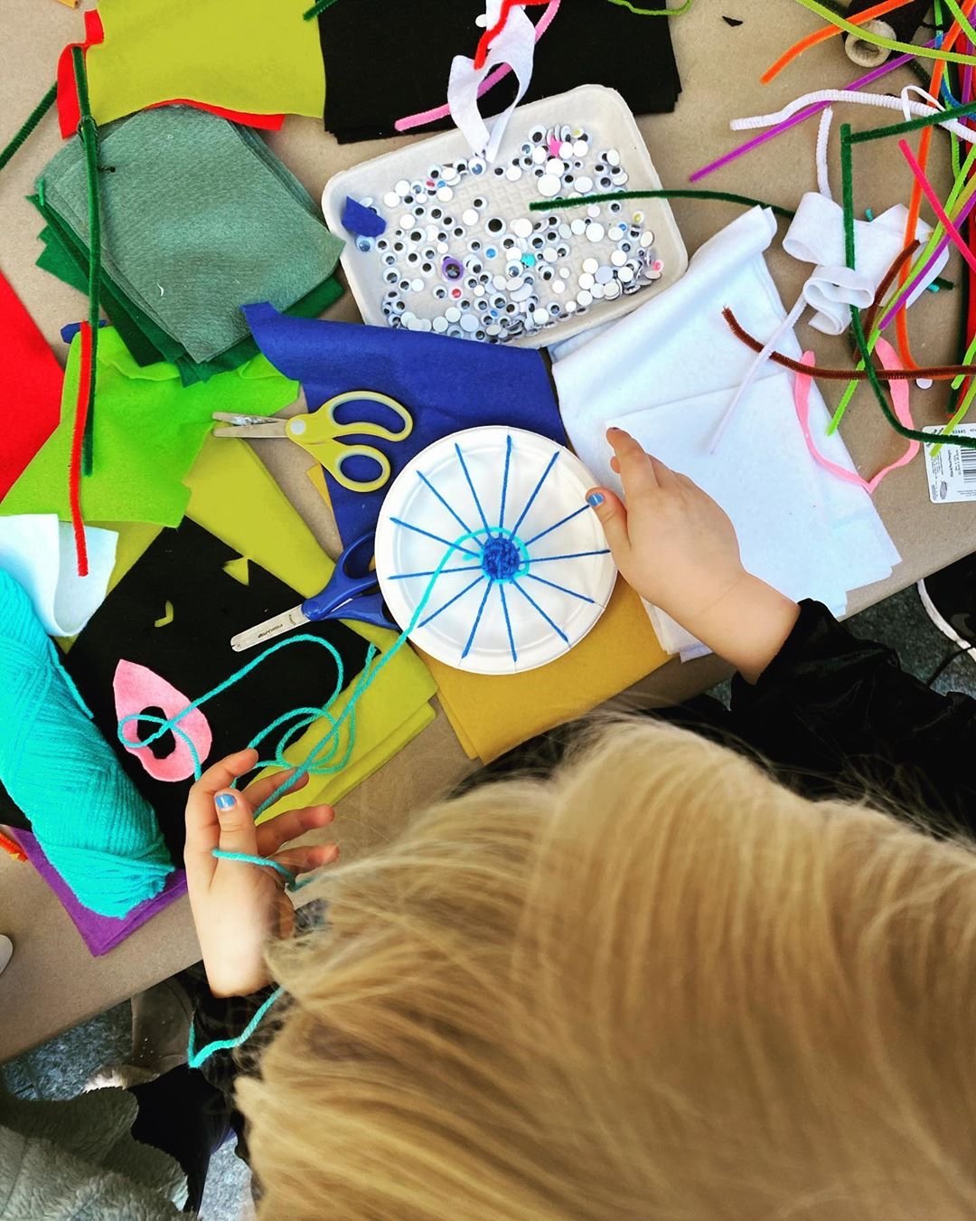 Child engaging in crafting activity with colorful felt, yarn, sequins, scissors, and paper plates in a craft station.