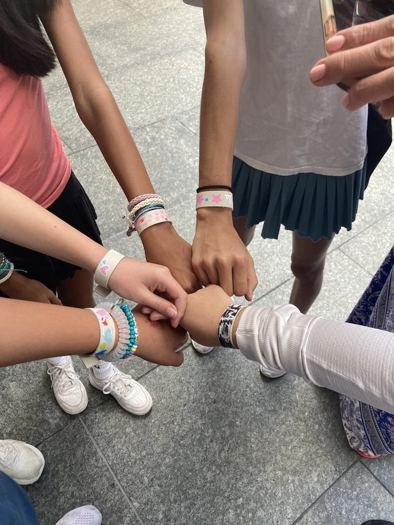 Six girls with various bracelets on their wrists, placing their hands together in a circle.