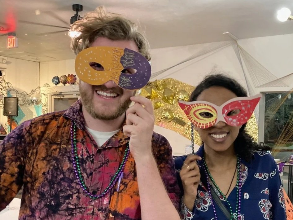 A man and a woman smiling behind handmade Mardi Gras masks at a Kiki Knows Art workshop in Dallas.