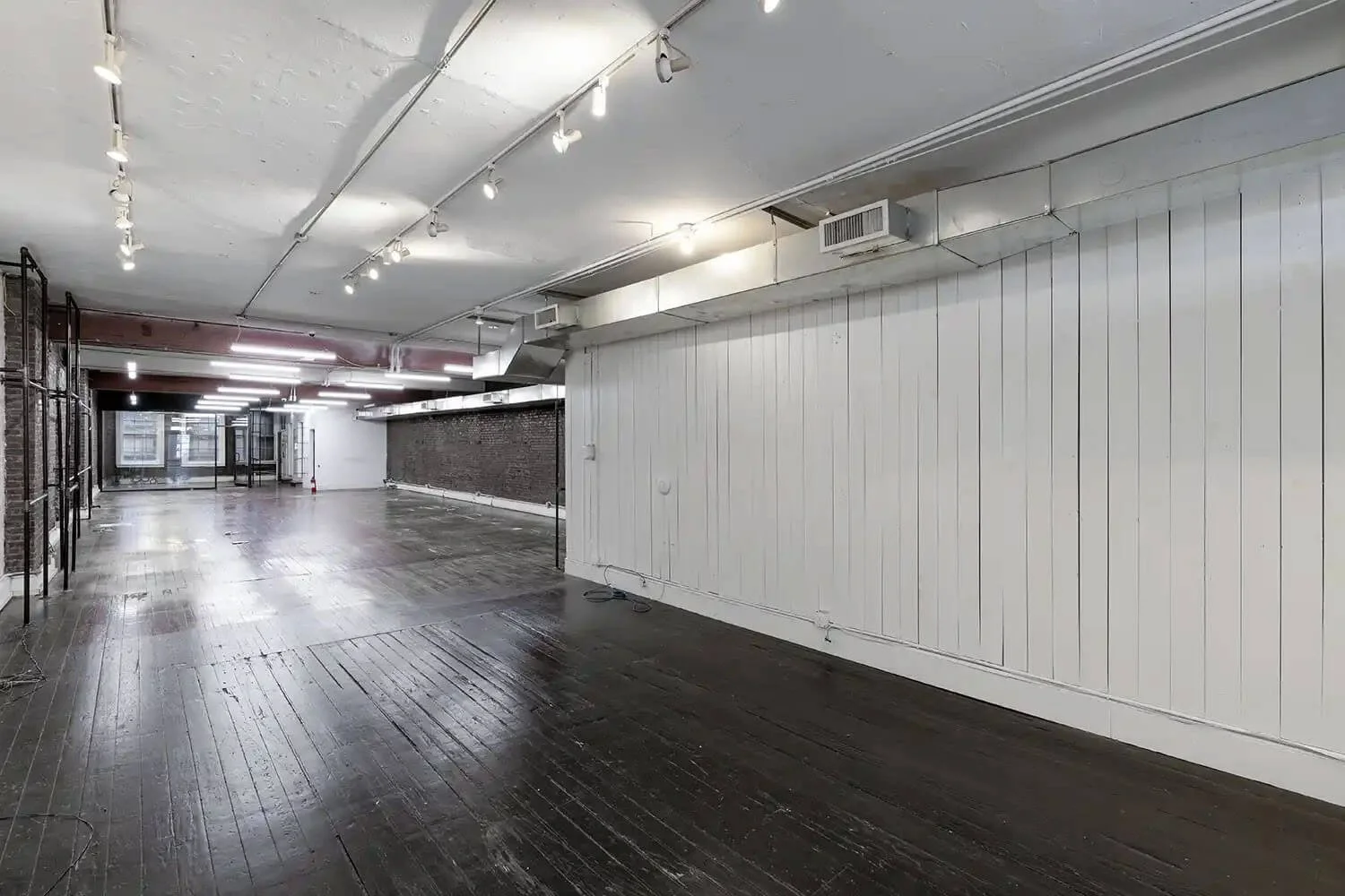 Empty industrial-style interior space with dark wooden floors, white paneled wall, exposed brick and metal стены, ceiling with track lighting, and large windows at the far end.