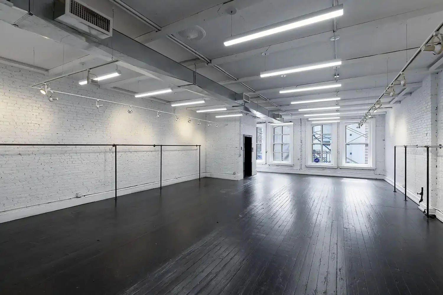 Empty art gallery with white brick walls, large windows, black wood floors, and ceiling tracks with lights.