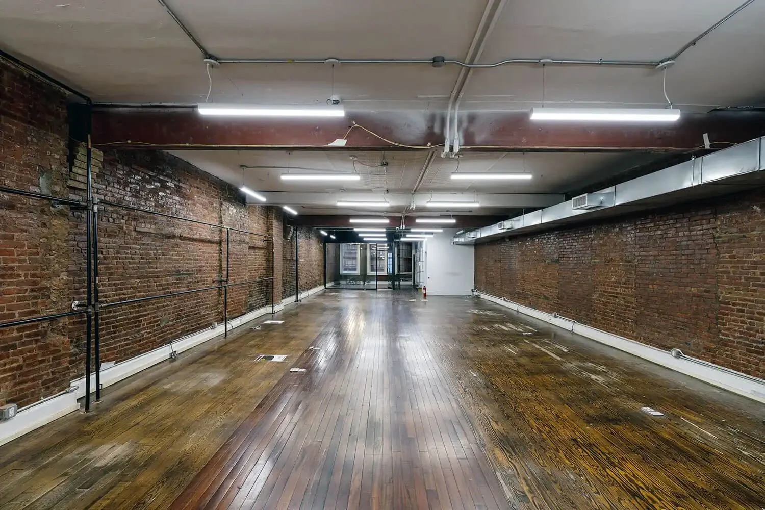 Empty industrial-style room with exposed brick walls, wooden floor, and ceiling lights.