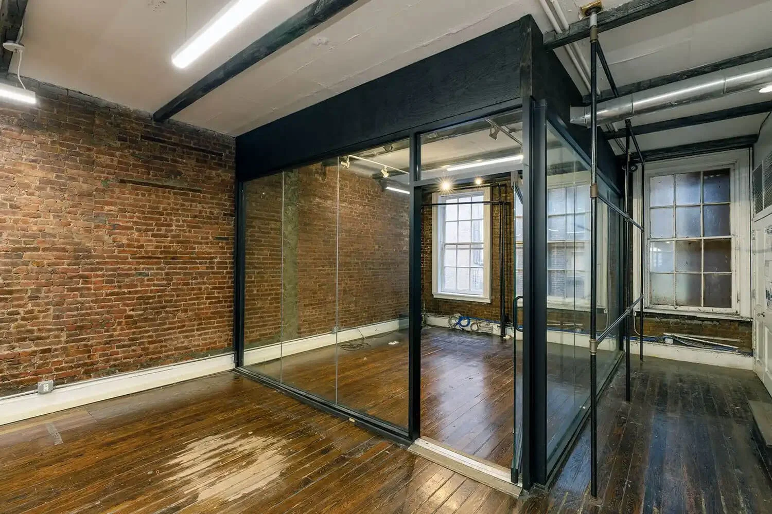 An empty industrial-style room with exposed brick walls, wooden flooring, large windows, and a glass enclosed office space with black metal framing.