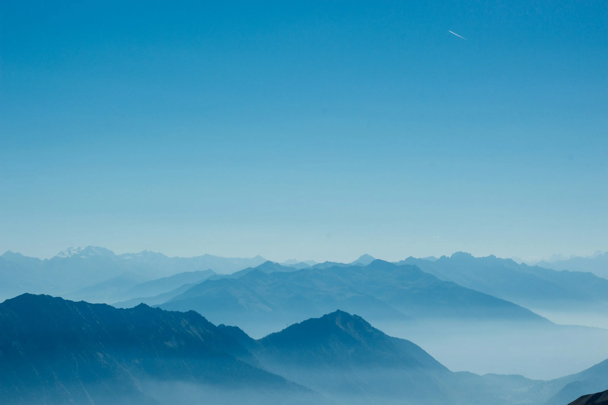 View of distant mountain range with snow-capped peaks under a clear blue sky, with a thin contrail visible.