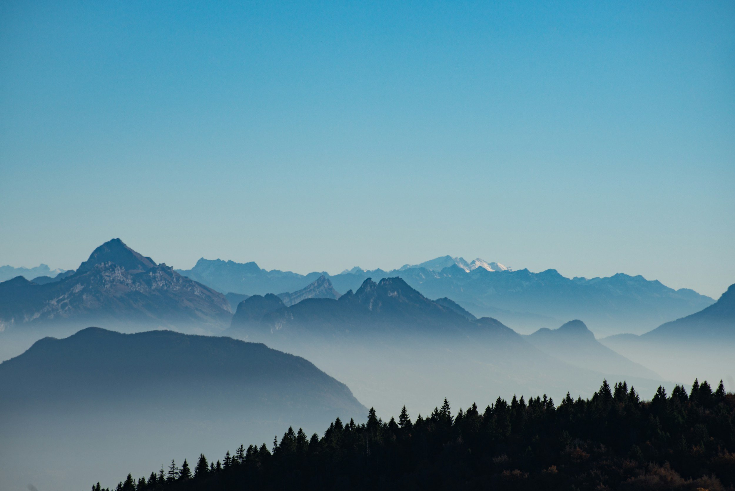 Mountain range with multiple peaks, layered with mist and snow, under a clear blue sky.