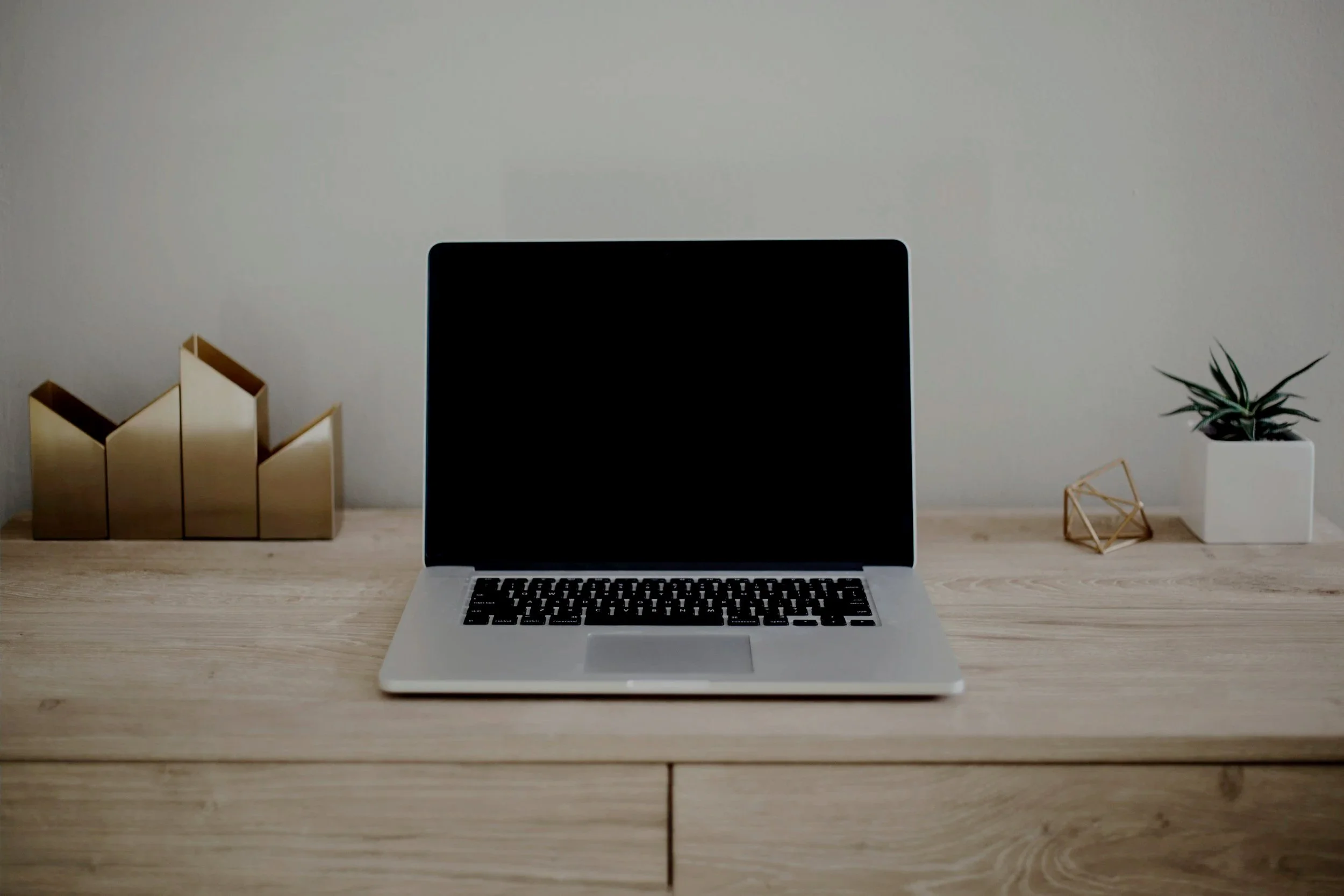 A laptop on a wooden desk, with a minimalist decor including a plant in a white pot, a gold geometric ornament, and several gold filing folders on the left side.