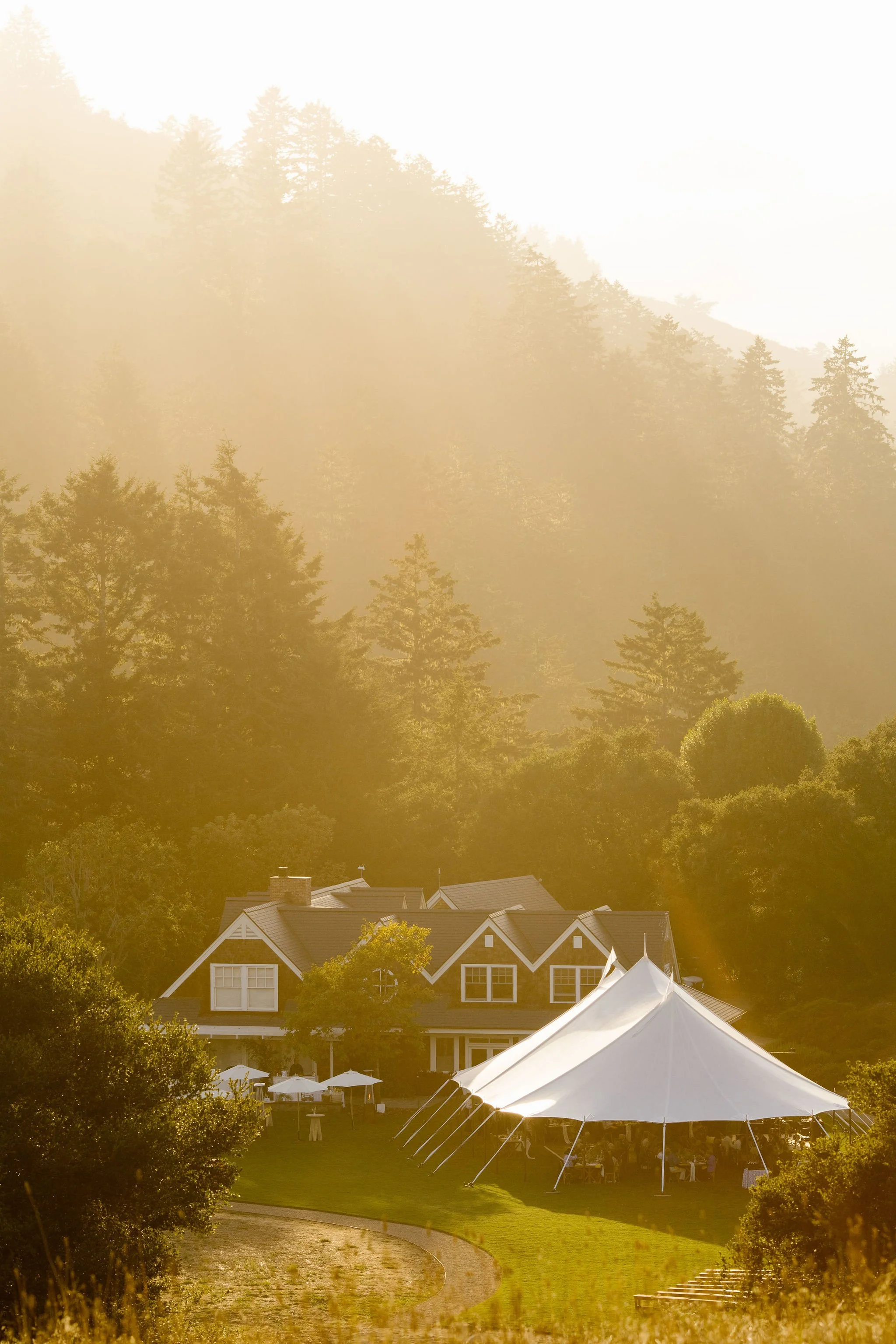 A large white event tent set up on a grassy area at the edge of a walking path. In the background, there is a two-story house with a dark roof and white window frames, surrounded by tall trees on a hillside. The scene is illuminated by golden sunlight