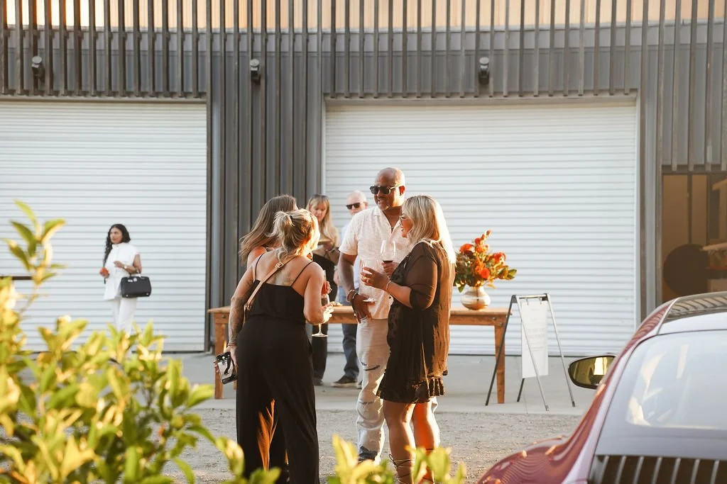 Group of people socializing outdoors near a building with large closed garage doors, with a table decorated with flowers and a man holding a drink, during sunset at a Porsche event.