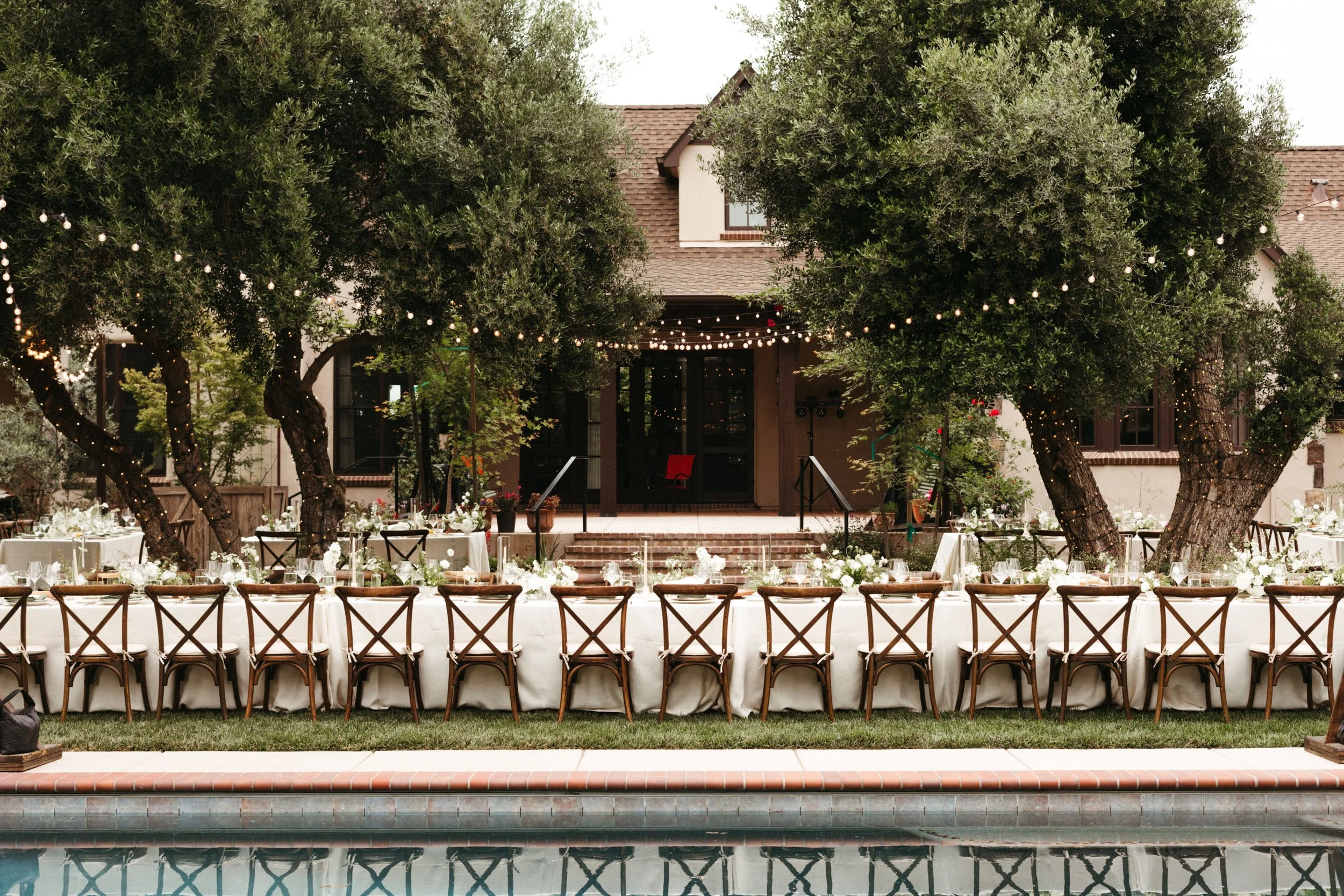 Wedding reception area set up outdoors with tables and chairs in front of a house, decorated for a celebration with string lights hanging from trees. Hosted in the Bay Area.