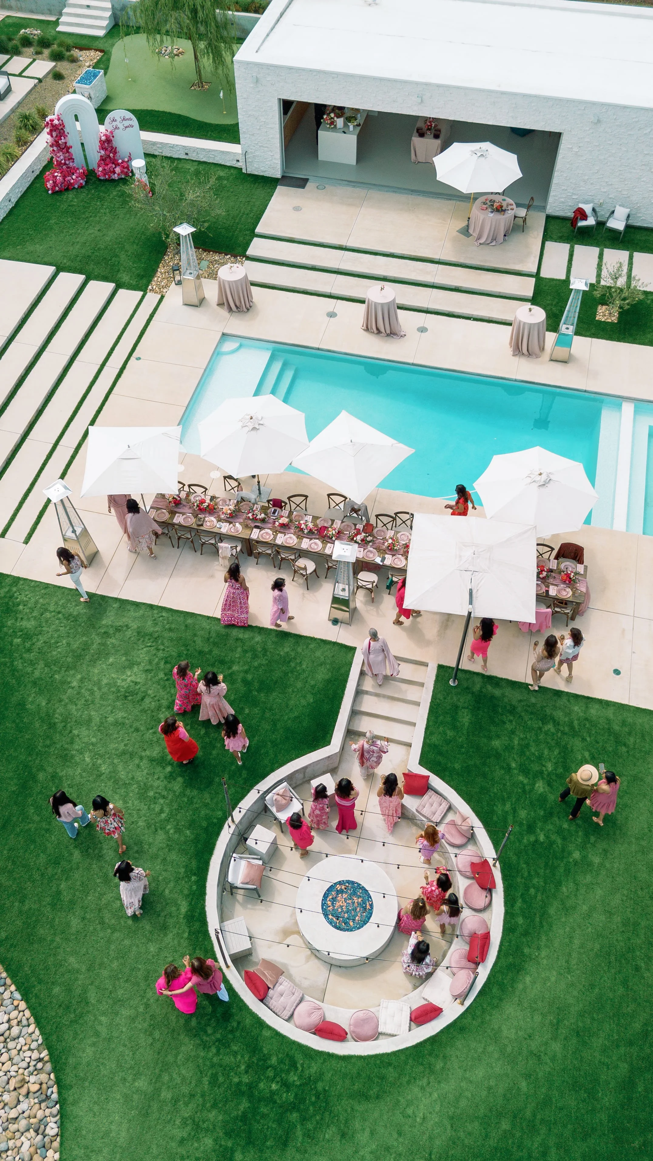 An aerial view of a backyard party produced by Urban Organics SF. Featuring pink decorations, a swimming pool, and outdoor furniture. People are gathered around tables and seating areas, with some children and adults mingling on the grass and patio.