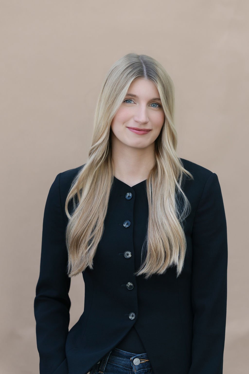 A young woman with long blonde hair, blue eyes, and light skin, wearing a black blazer and dark jeans, standing against a plain beige background.