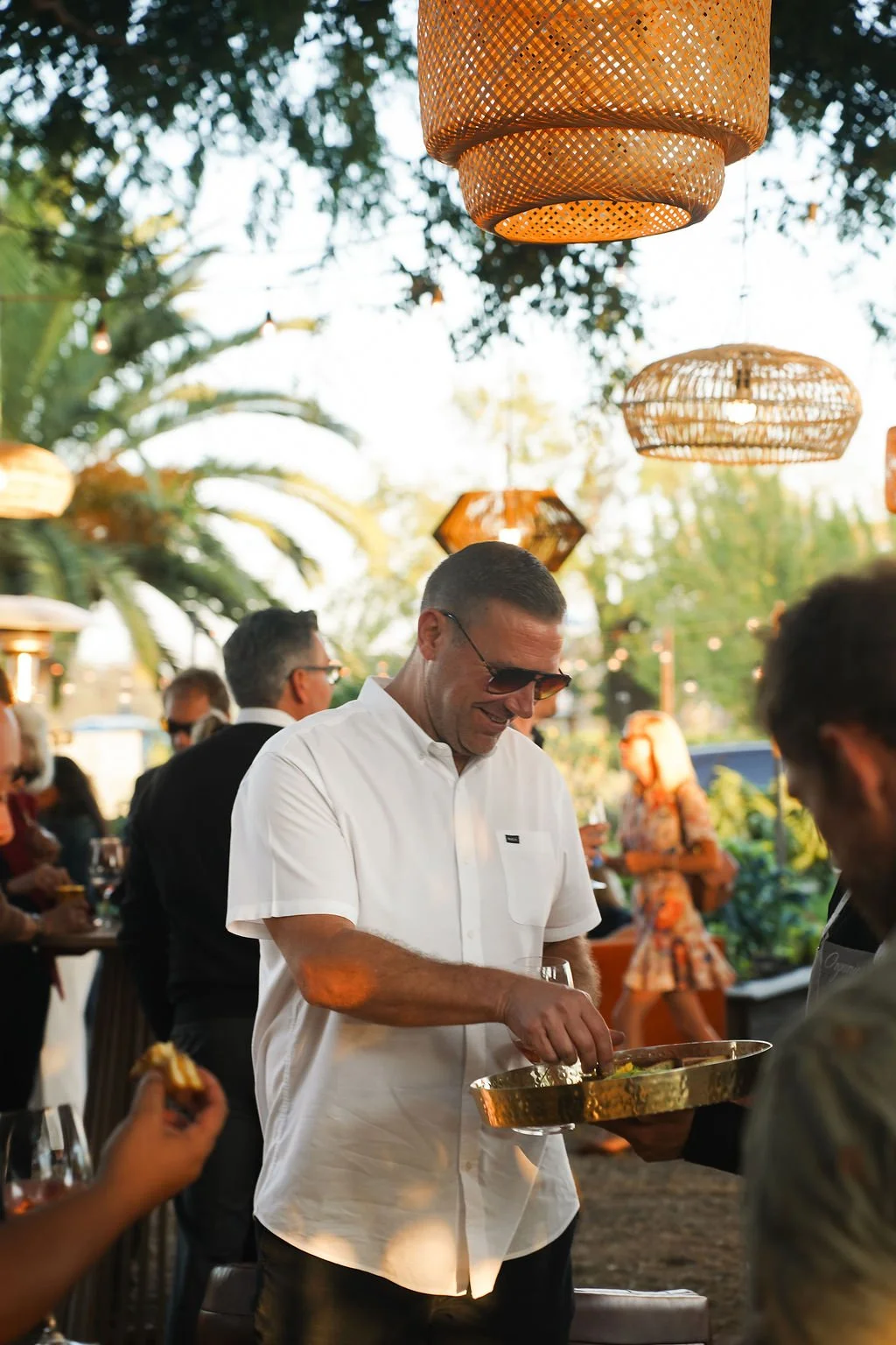 Porsche corporate event with a man in a white shirt and sunglasses at an outdoor gathering, serving food from a tray, with hanging wicker lanterns and other guests in the background.