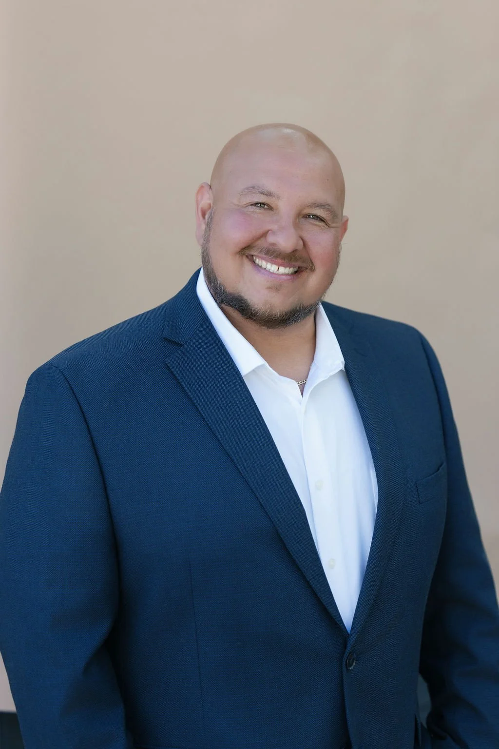 A smiling man in a navy blue suit with a white shirt standing against a plain beige wall.