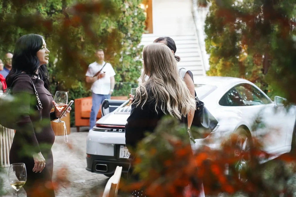 Porsche event, socializing outdoors near a white sports car, with some holding drinks and others engaging in conversation, surrounded by foliage.