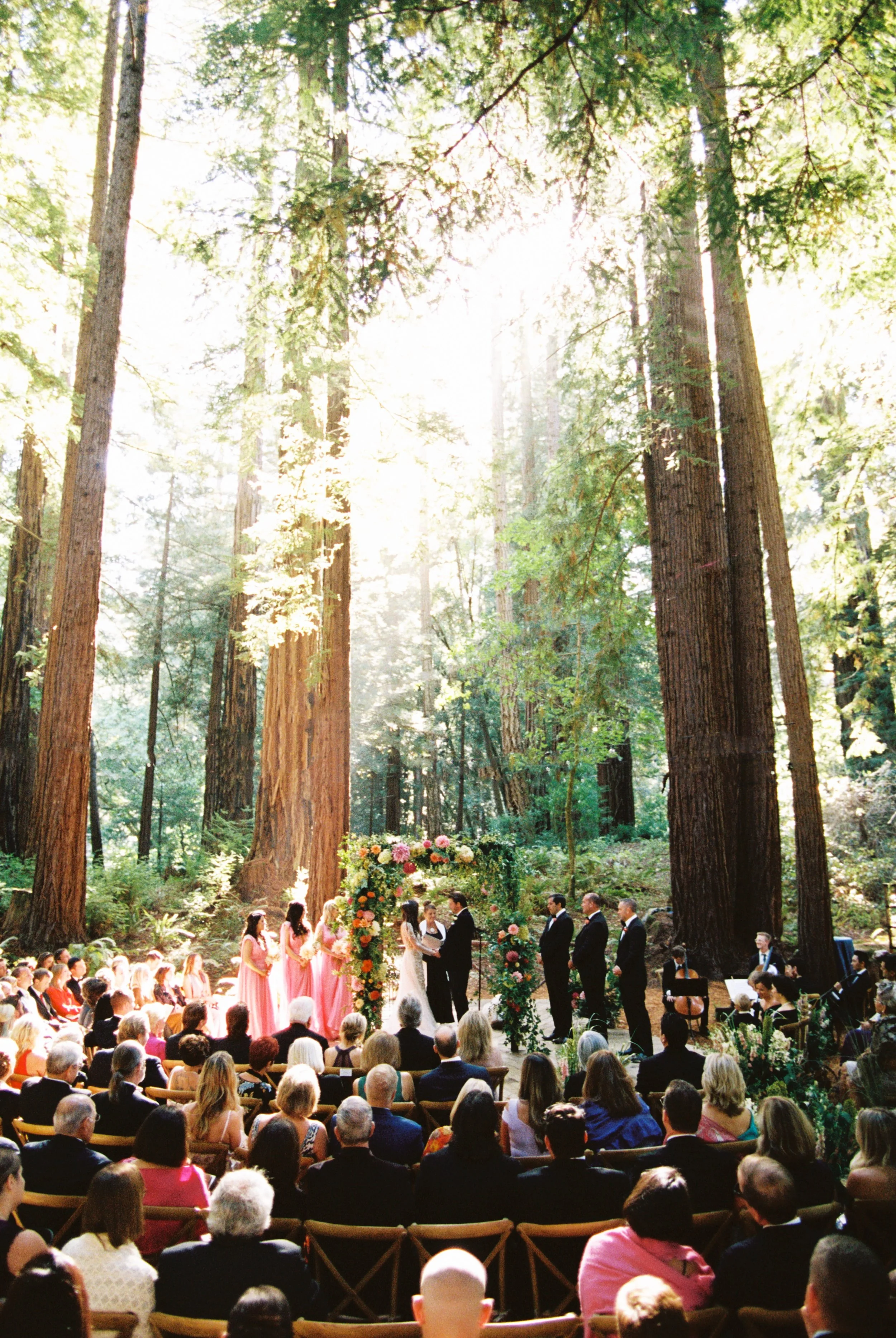 An outdoor wedding ceremony in a forest with tall trees and sunlight shining through the leaves. Guests are seated watching the couple exchange vows at an altar decorated with flowers in San Francisco.