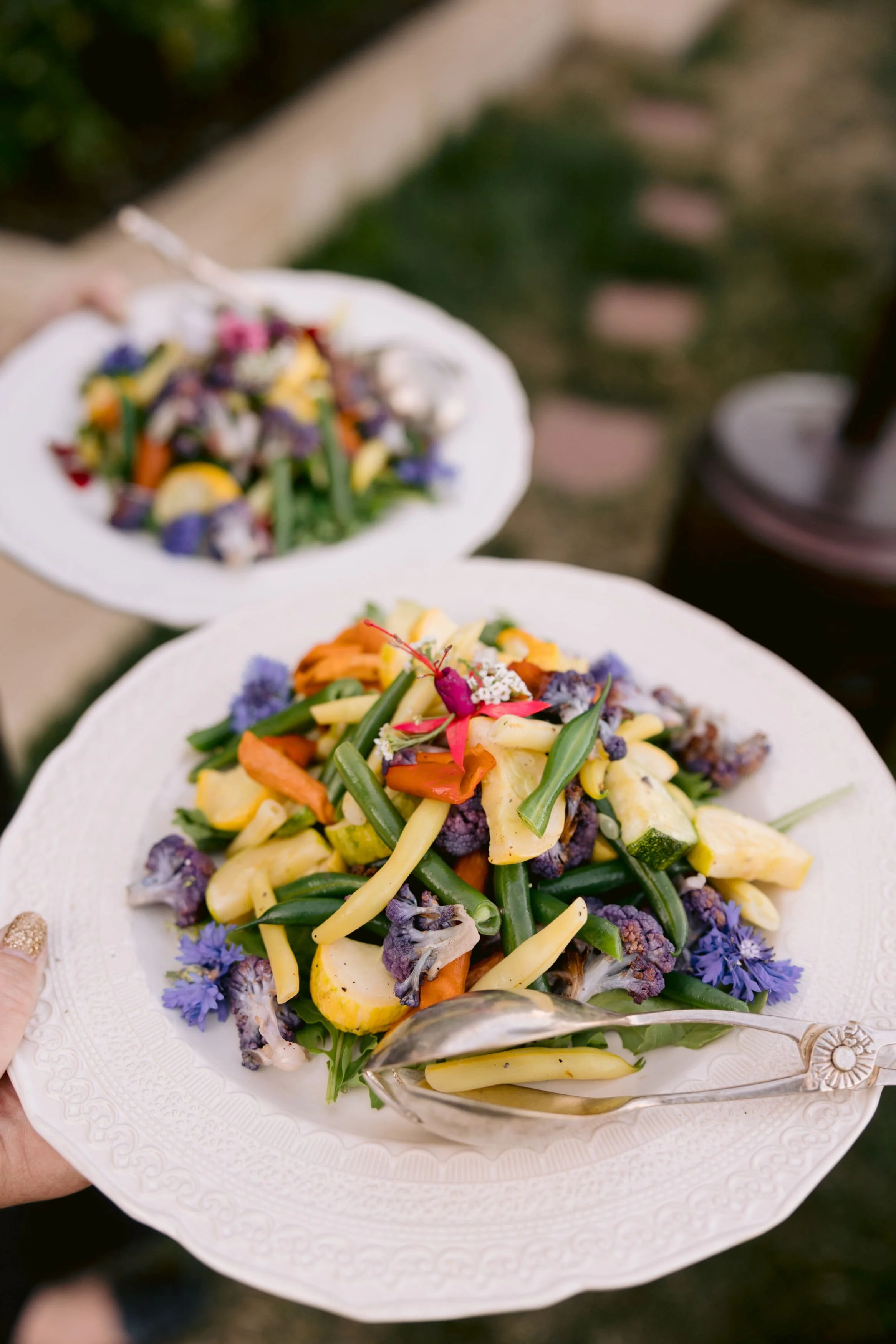 Corporate event with a white decorative plate holds a colorful salad with various vegetables including green beans, carrots, and purple cauliflower, garnished with edible flowers and herbs, held outdoors.