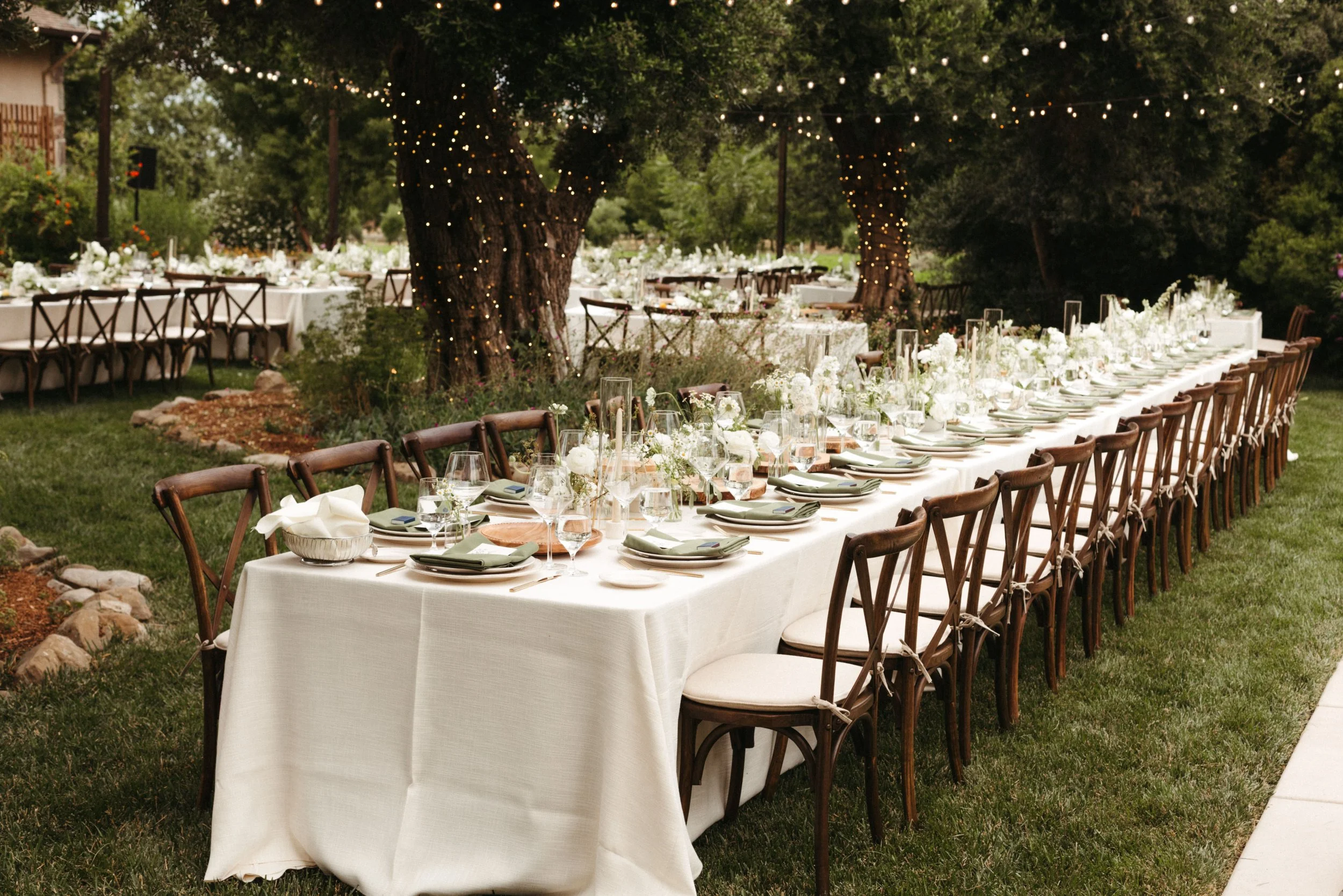 Outdoor Bay Area wedding reception setup with long dining tables covered in white tablecloths, decorated with floral arrangements, surrounded by wooden chairs, under large trees adorned with string lights during evening.