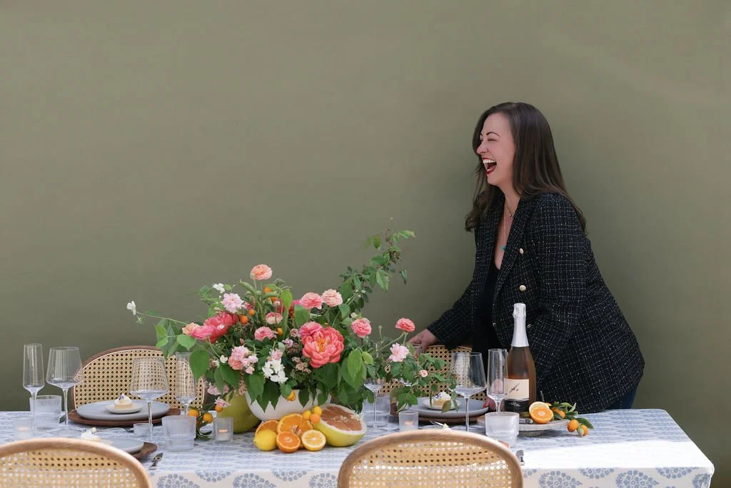 Brittany Maffia CEO and Founder of Urban Organics laughing at a decorated dining table with a large floral centerpiece, plates, glasses, and a bottle of champagne.