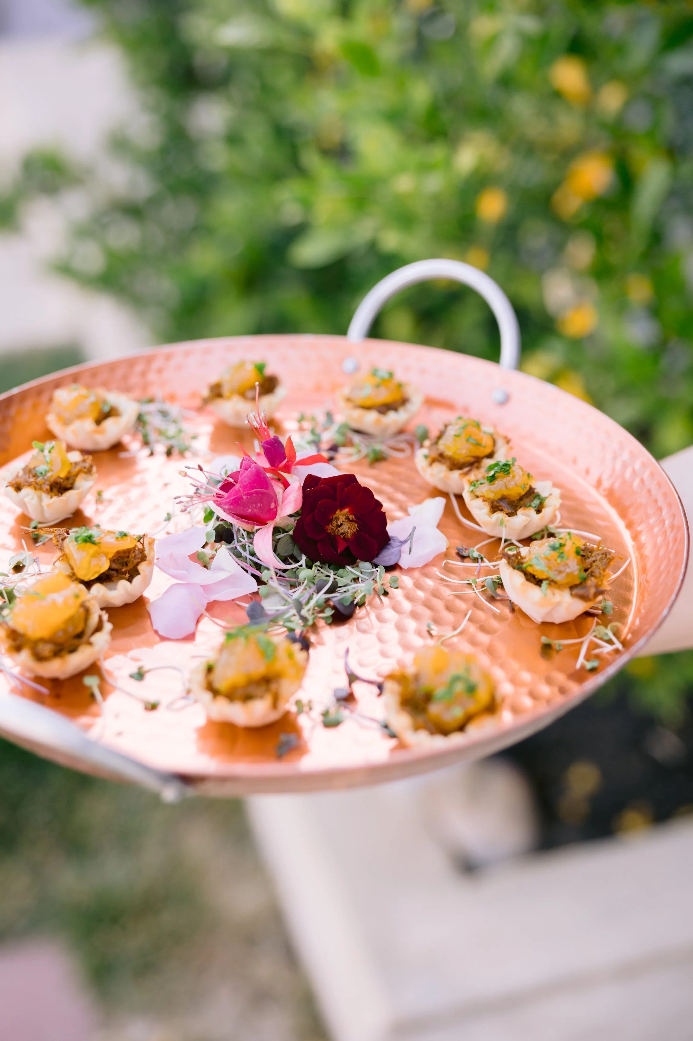 A round metal tray with small seafood appetizers, garnished with flowers and herbs, outdoors with blurred green foliage in the background. Culinary experience designed by Urban Organics.
