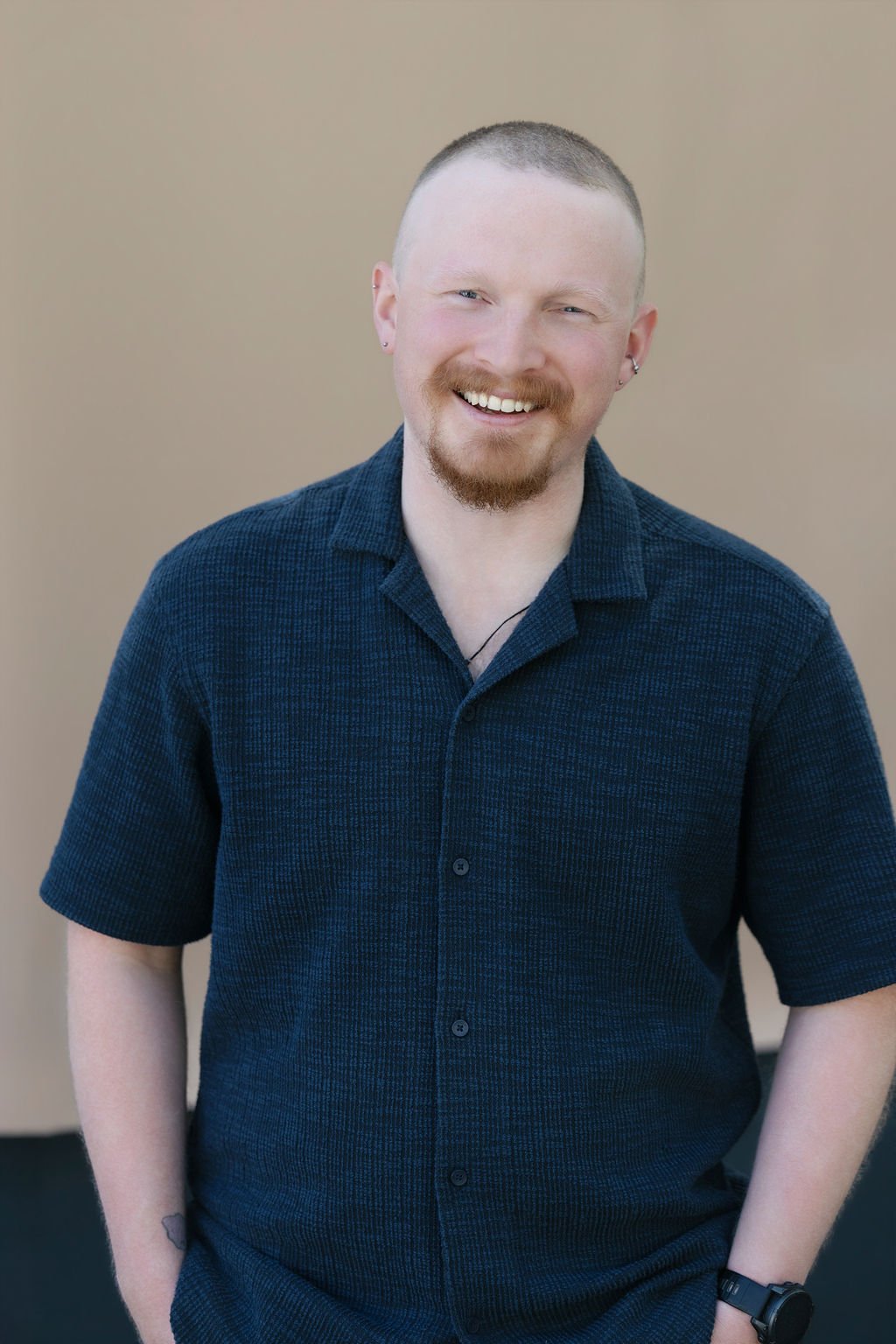 A young man with short blonde hair and a beard, smiling, wearing a dark blue short-sleeved button-up shirt, standing against a plain beige background.