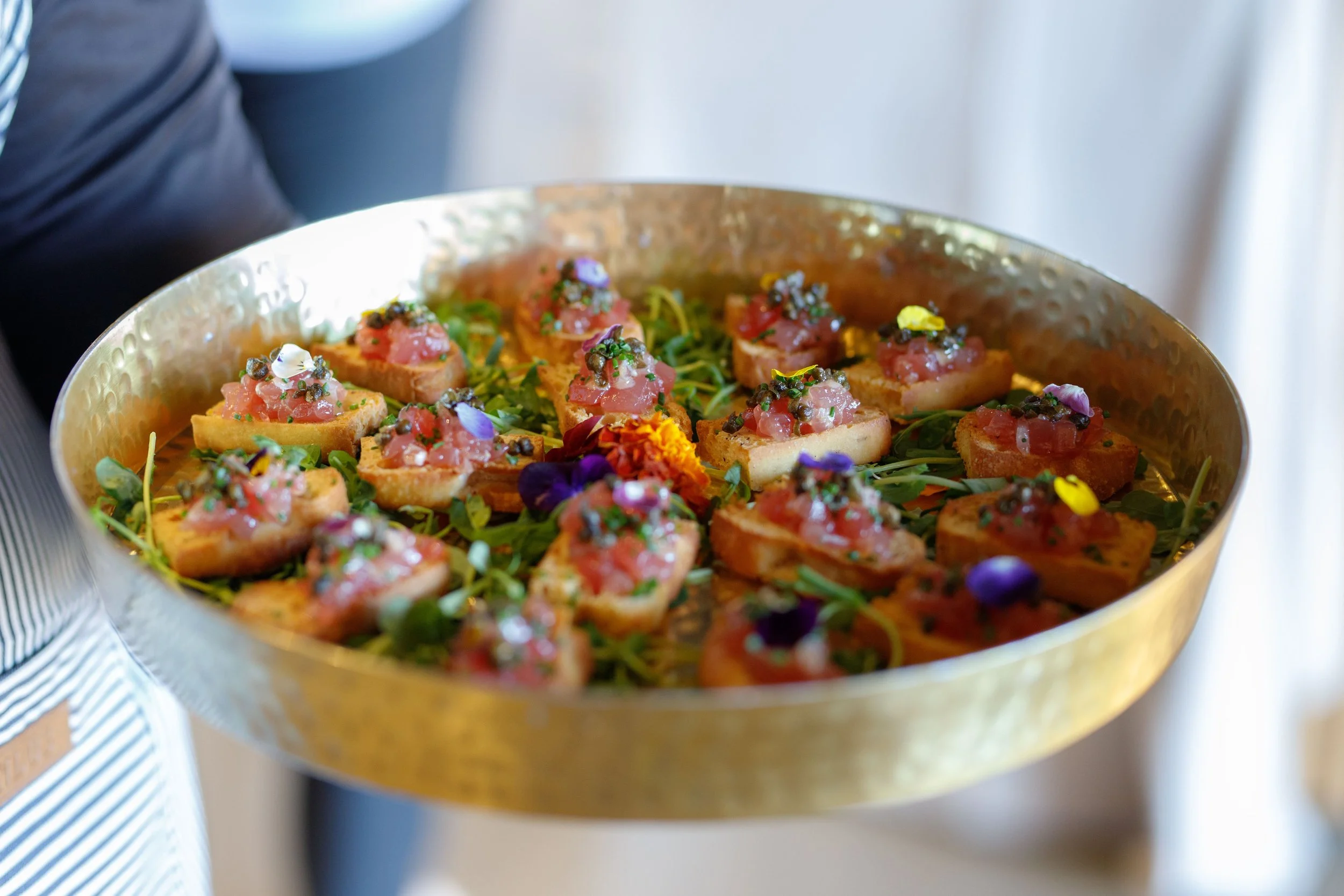 Culinary service featuring a golden tray containing small pieces of toast topped with diced raw fish, garnished with colorful edible flowers and microgreens, likely an appetizer or canapés.