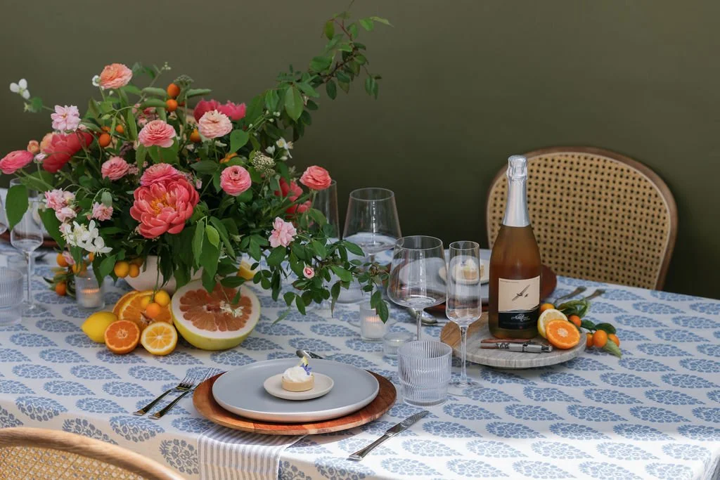A dining table decorated with a large floral arrangement, sliced citrus fruits, a bottle of sparkling wine, and various glasses, set with plates, silverware, and a woven chair in the background. Design by Urban Organics SF