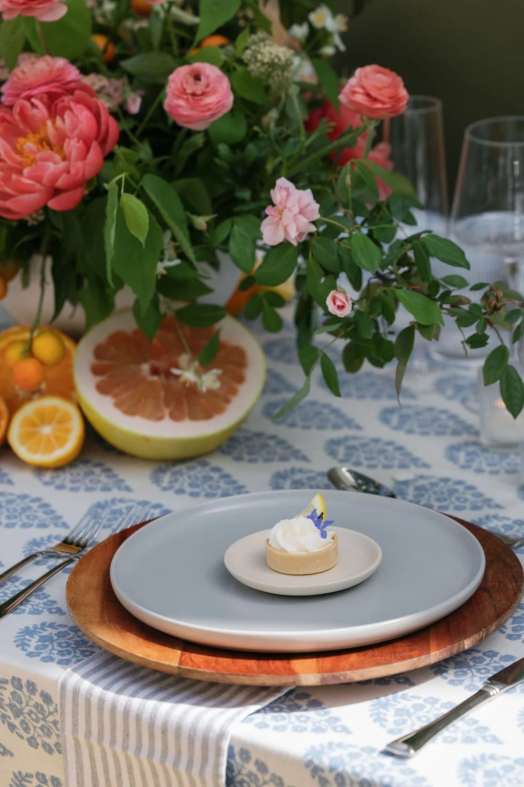 An elegantly set dining table with a floral centerpiece, slices of citrus fruits, and a dessert on a plate. The table features a blue and white patterned tablecloth, and there are empty wine glasses in the background in Bay Area.