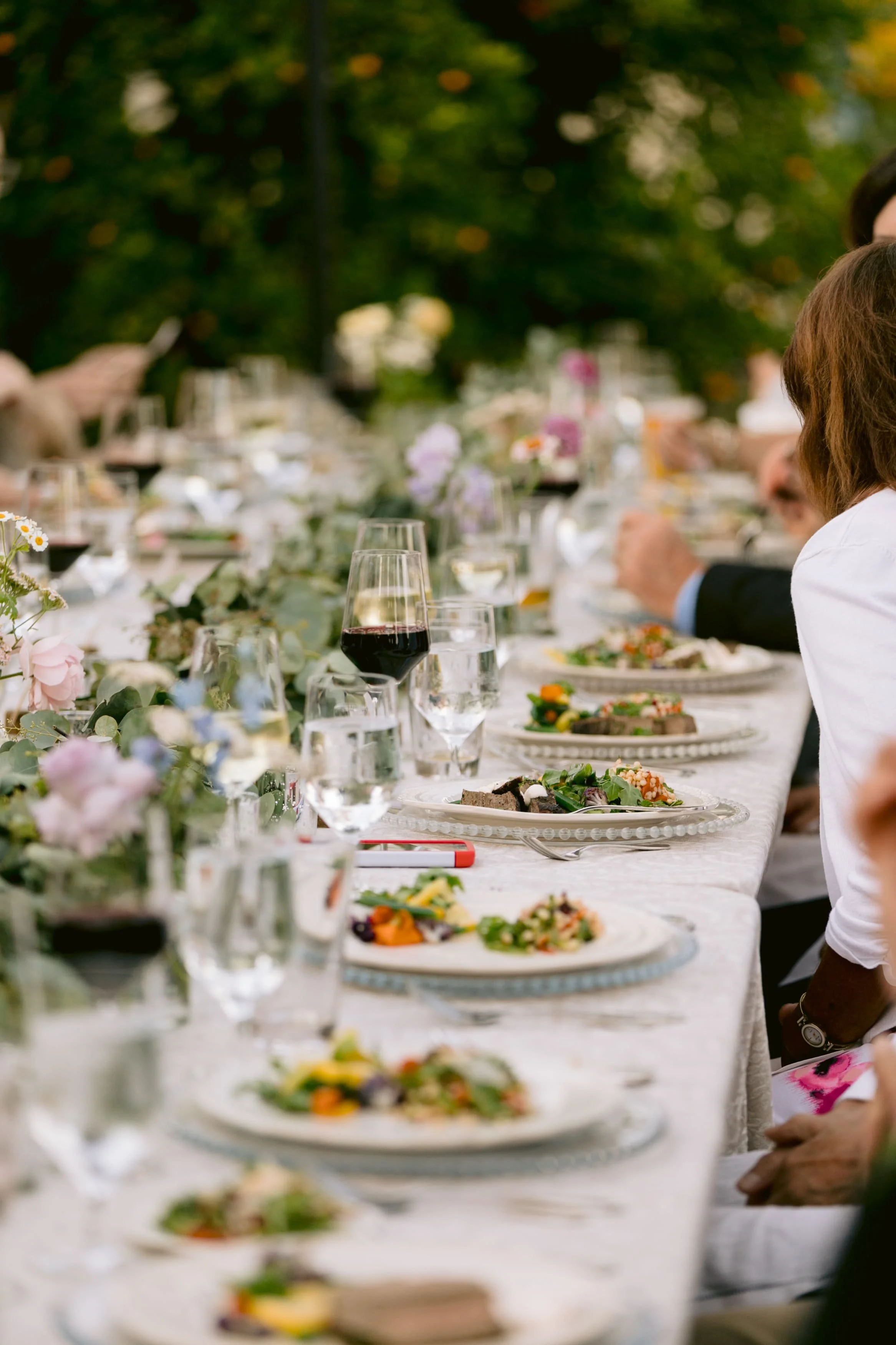 A long outdoor dining table with plates of food, glasses of wine, and floral centerpieces, set in a garden with trees in the background. Produced by Urban Organics in the Bay Area.
