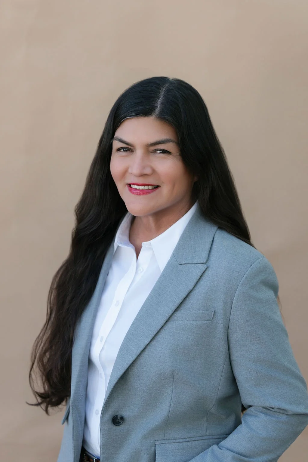 Professional woman with long dark hair wearing a gray blazer and white shirt, smiling, against a neutral background.