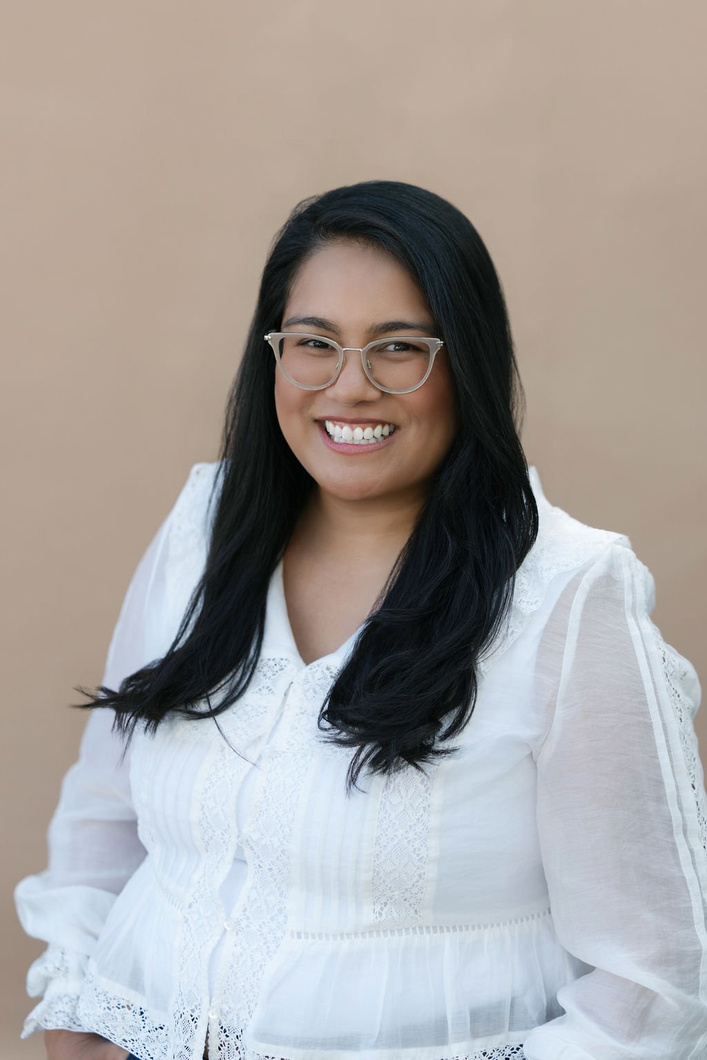 A woman with long black hair and glasses smiling at the camera, wearing a white blouse with lace details, standing against a beige background.