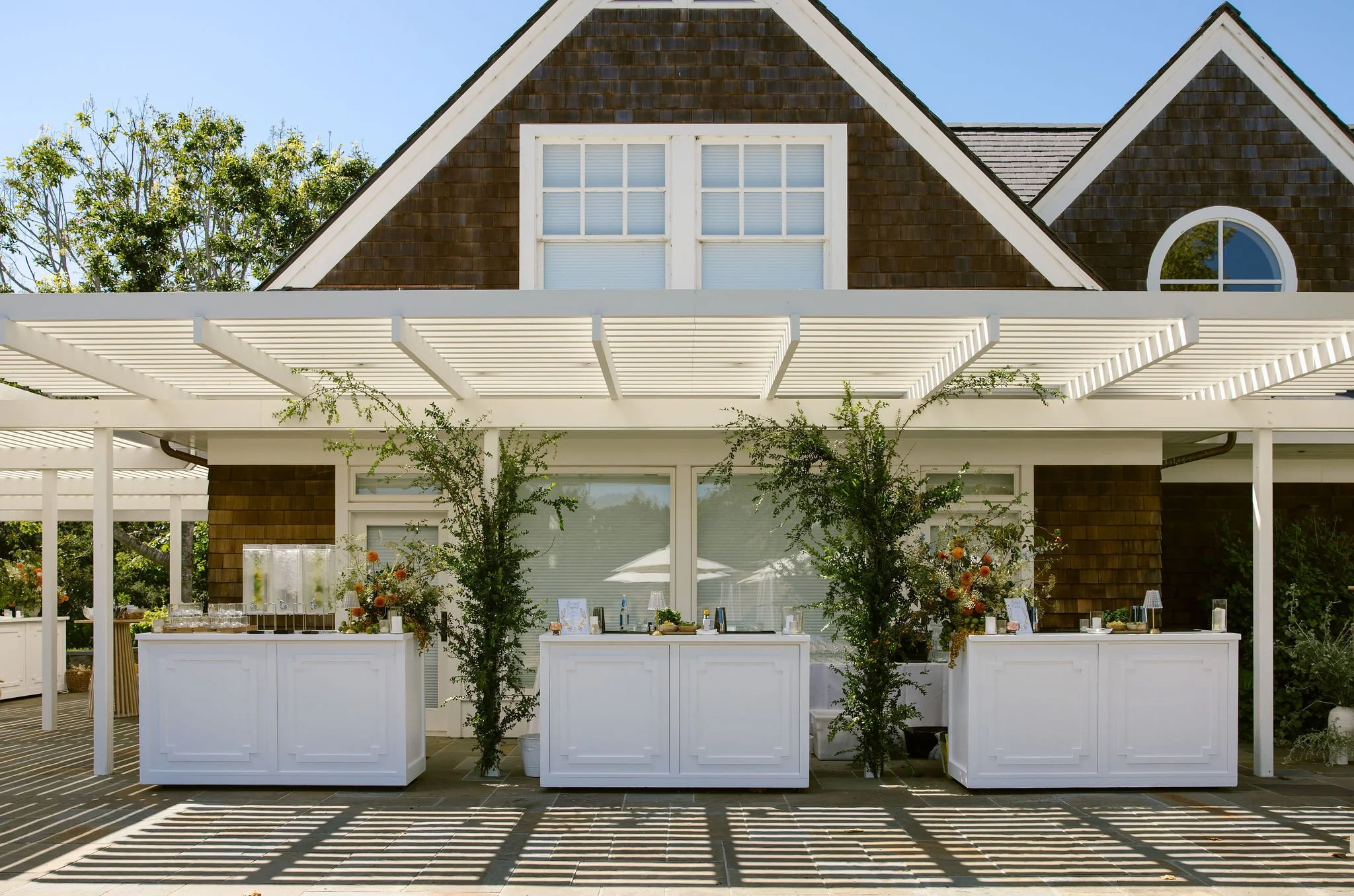 Outdoor wedding reception setup with white tables, floral arrangements, and a white pergola in front of a house with a brown shingle roof and multiple window styles, including a large rectangular window and a round window, under a clear blue sky.