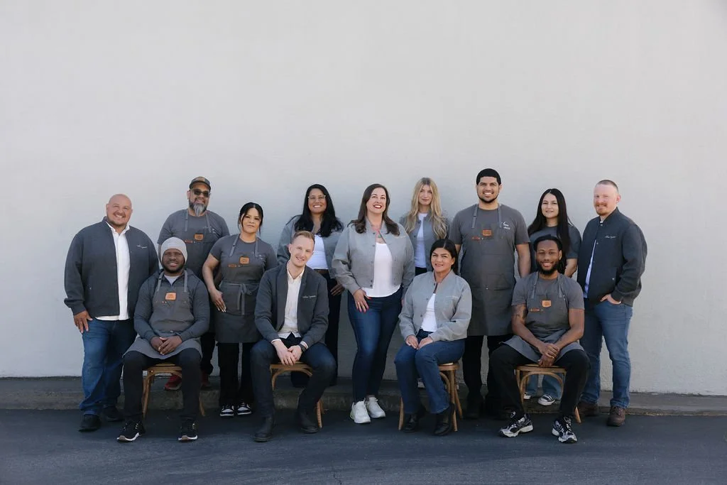Group of twelve people from the Urban Organics team, some seated and some standing, posing outdoors against a plain wall, with some wearing gray uniforms.