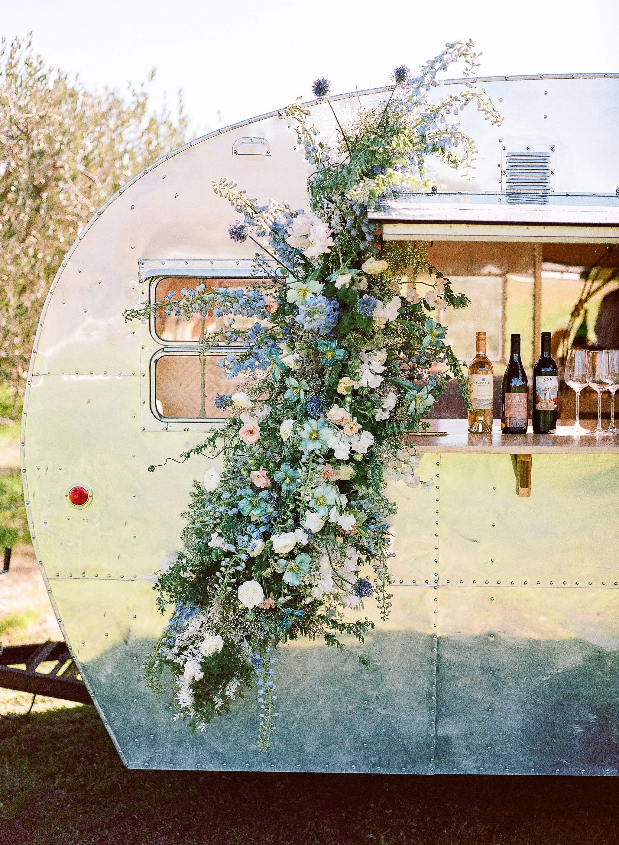 A vintage silver trailer decorated for a private event with a floral arrangement of white, blue, and purple flowers hanging down the side, with a small window and bottles of wine inside. Curated private dining experience.