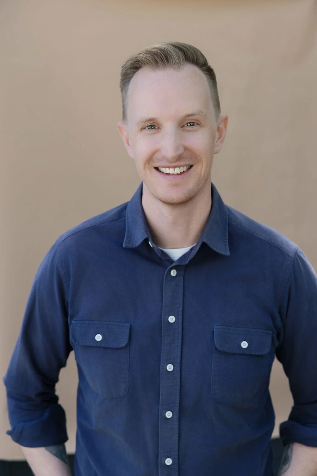 A young man with short blonde hair, blue eyes, and fair skin, smiling, wearing a blue button-up shirt with rolled-up sleeves, standing outdoors against a light brown background.