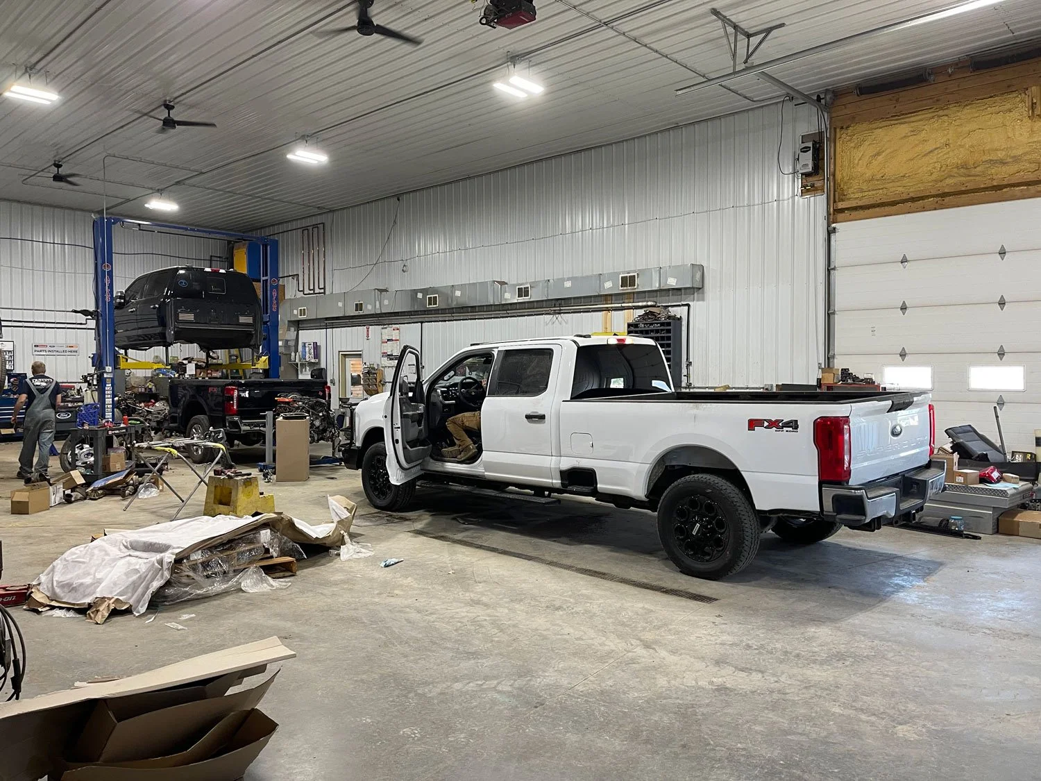 Inside an auto repair shop with a white pickup truck in the foreground and a black vehicle on a lift in the background. Cluttered workspace with tools, boxes, and equipment scattered around.