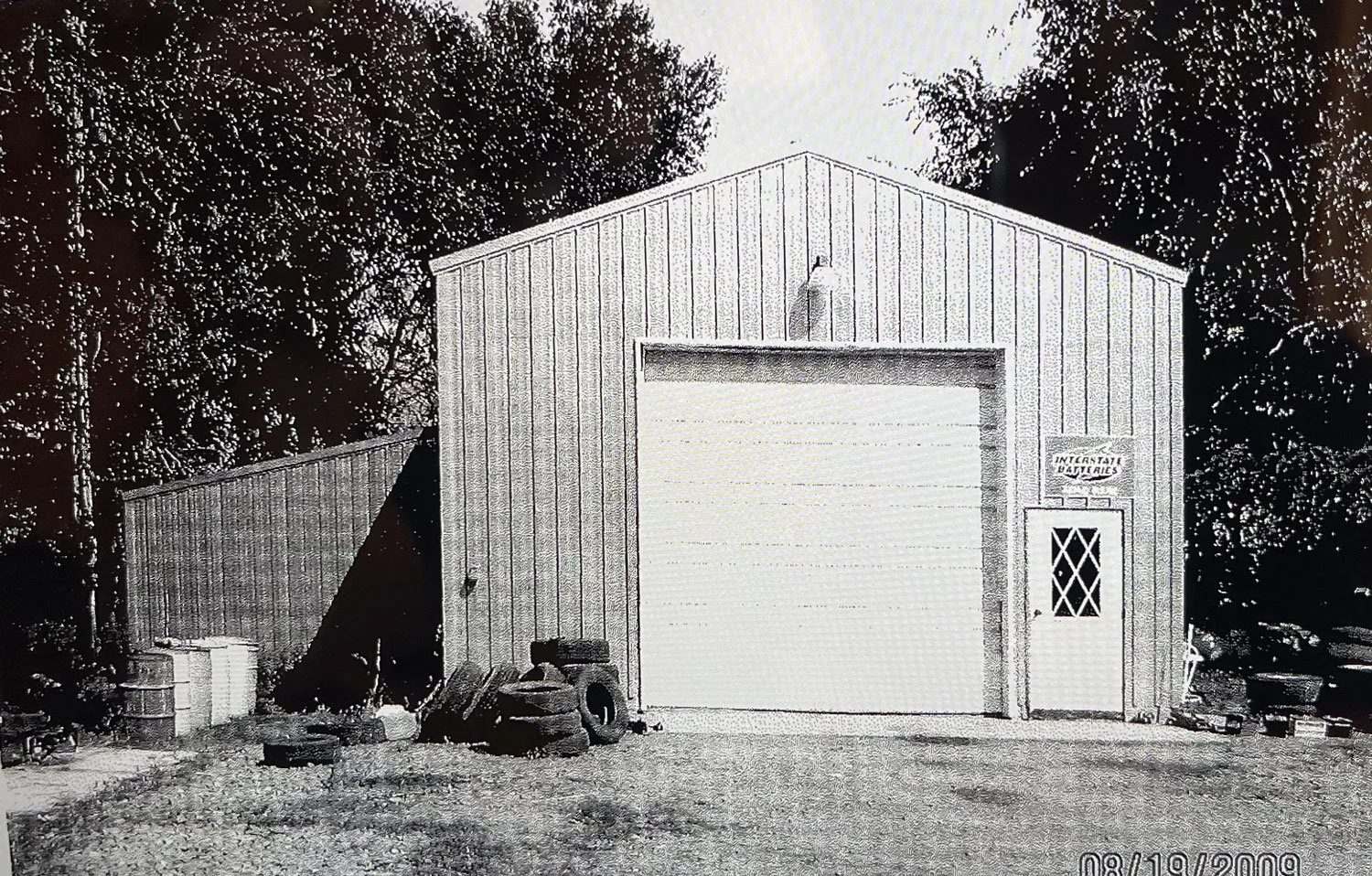 A metal building with a roll-up garage door and a smaller door to the right, located in an outdoor area with trees in the background. There are tires and miscellaneous items outside the building.