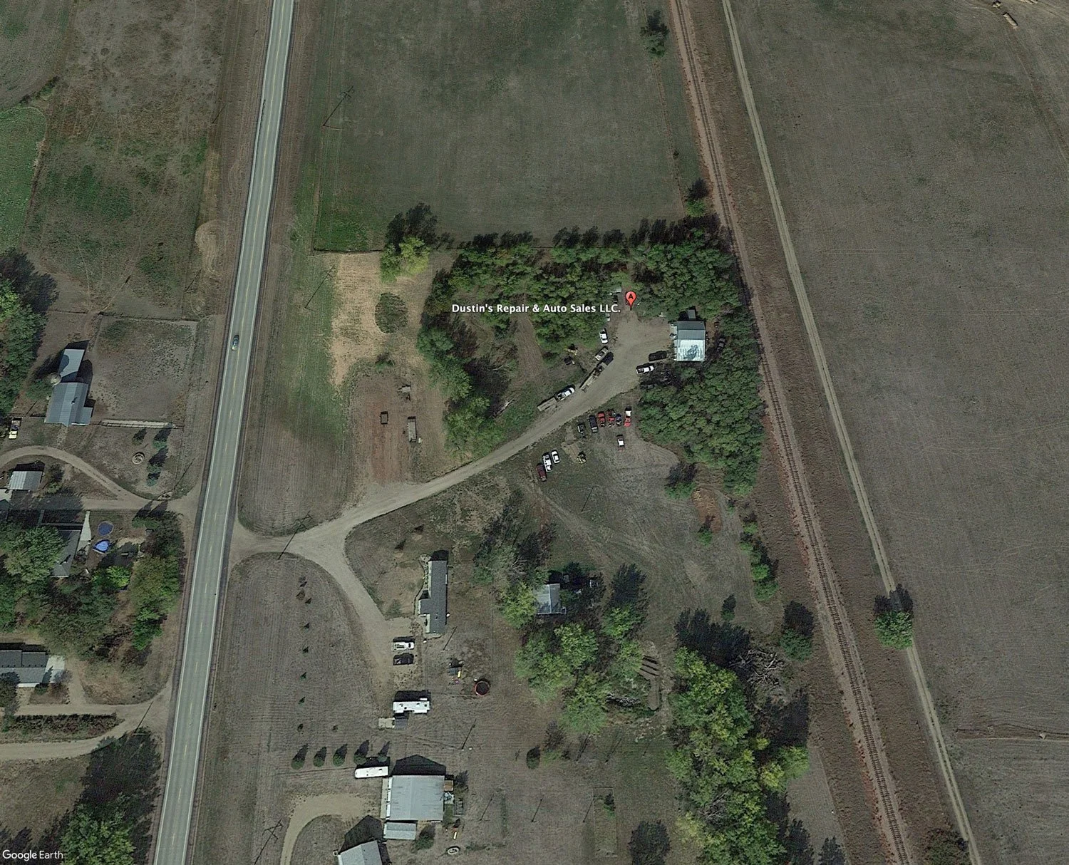 Aerial view of Dusty's Repair & Auto Sales LLC, with a parking lot, surrounding trees, and nearby fields, adjacent to a highway and railroad tracks.