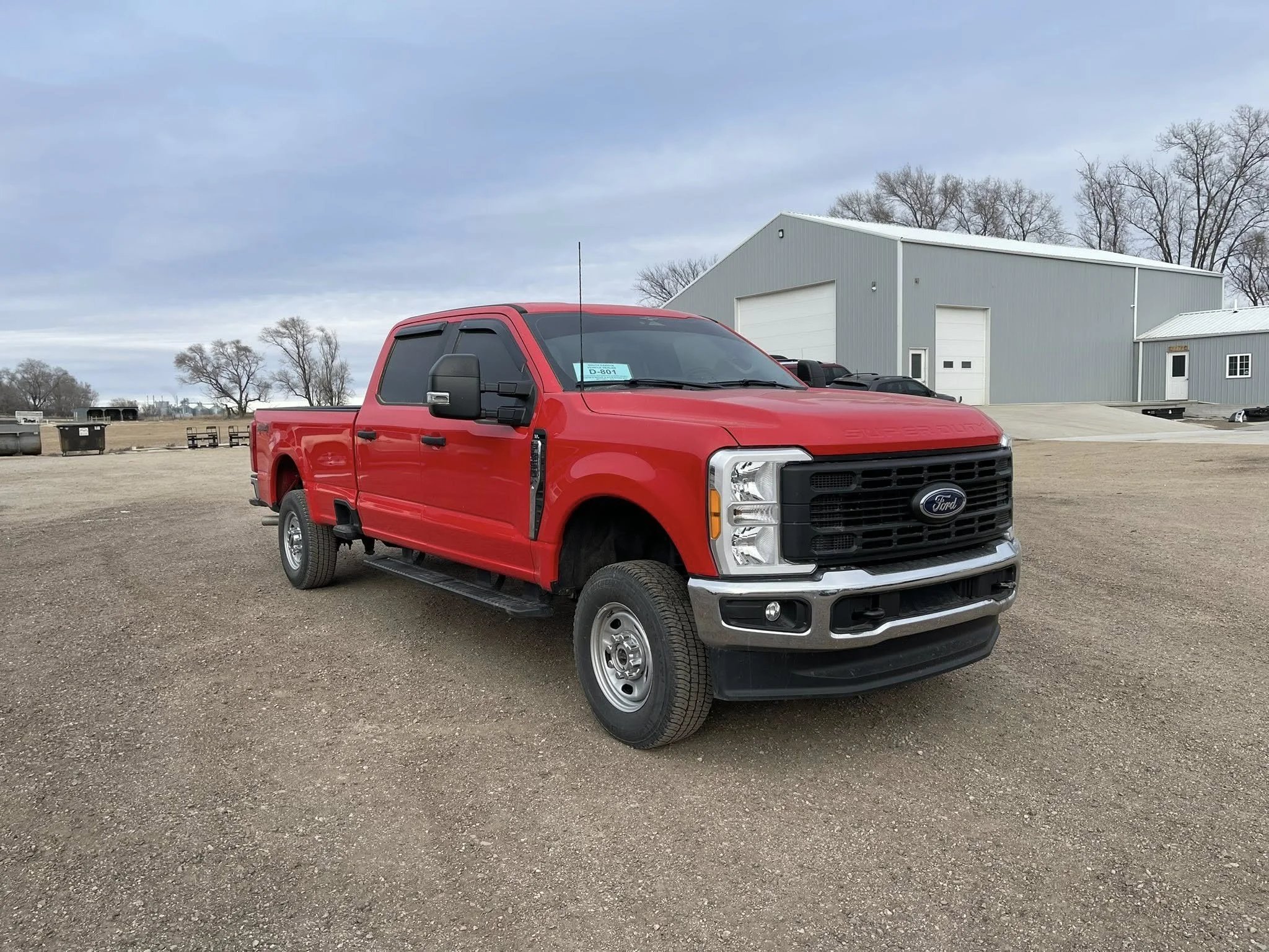Red Ford pickup truck parked on dirt lot near a gray industrial building with trees in the background.