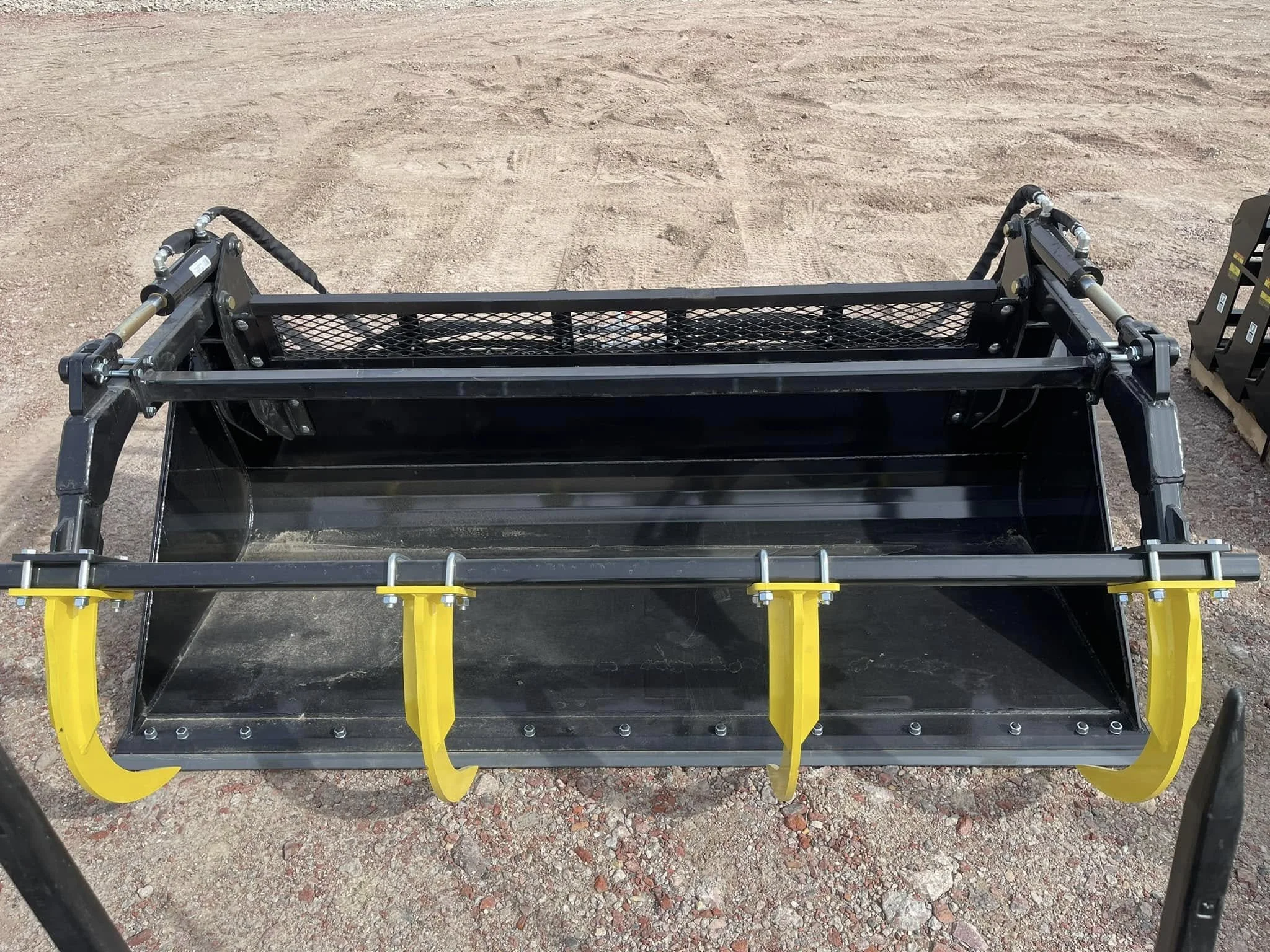 A front view of a construction machine attachment with black metal frame, yellow safety guard, and hydraulic cylinders, on a dirt surface.