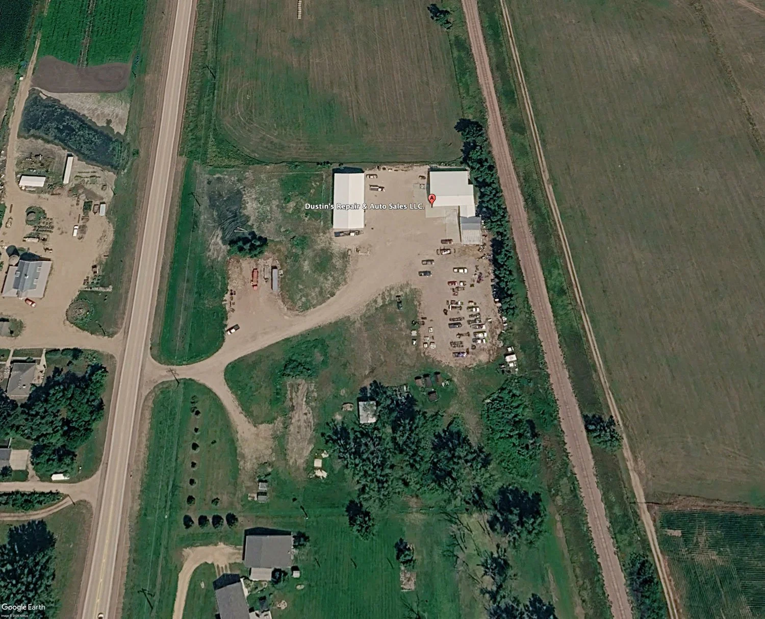 Aerial view of a rural property with a building labeled 'Dustin's Repair & Auto Sales LLC', surrounded by a parking lot, trees, and farmland.