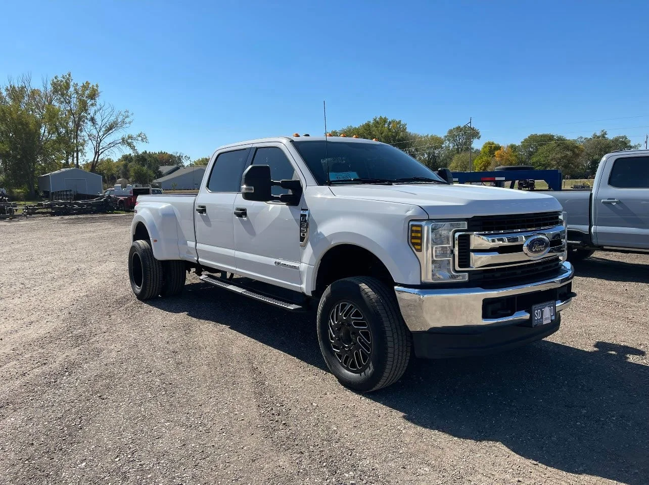 White Ford F-150 pickup truck parked on a gravel lot with trees and other vehicles in the background under a clear blue sky.